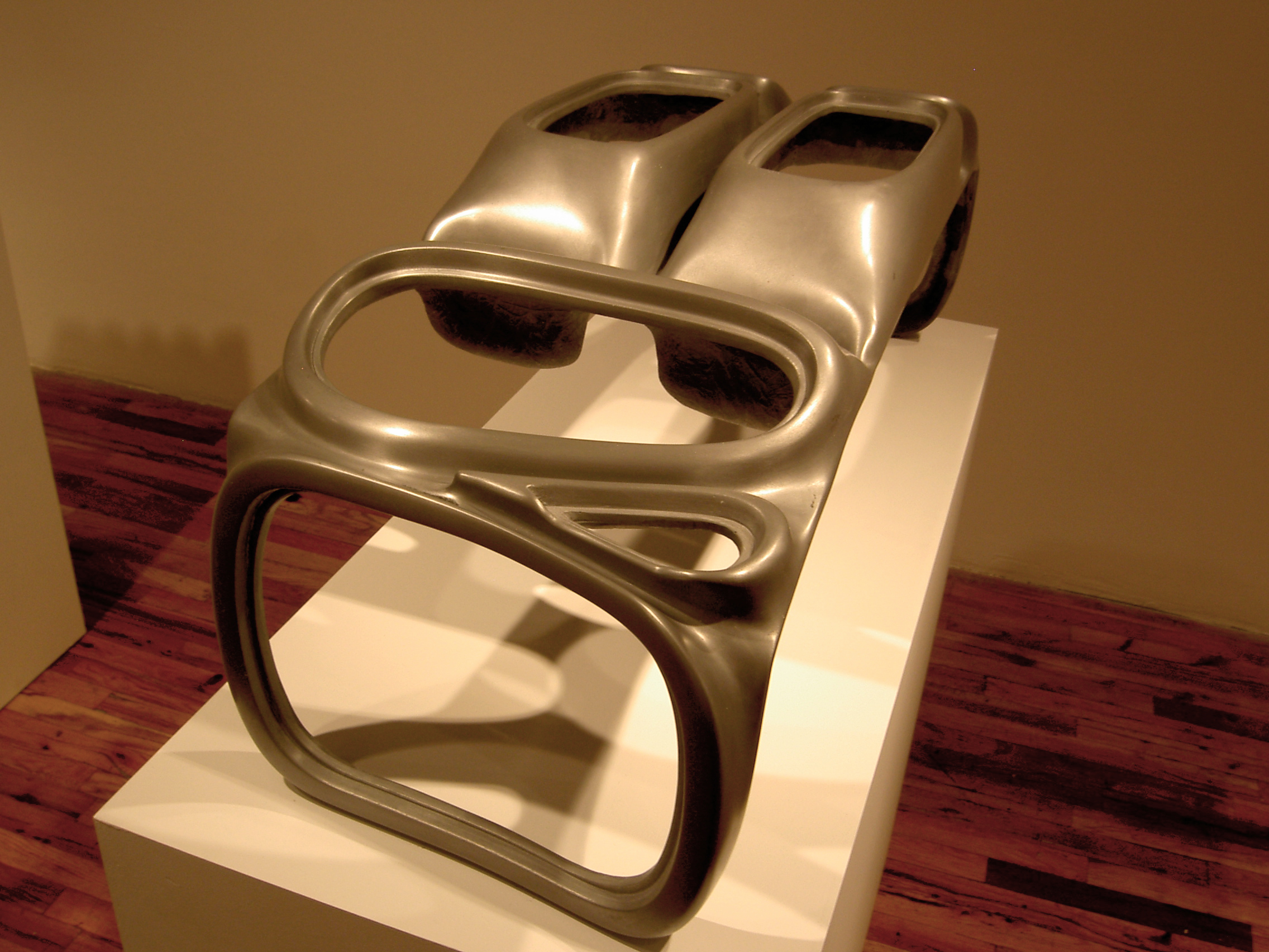 An abstract sculpture with a chrome finish is displayed on a white pedestal. The sculpture's form includes amorphous voids alongside somewhat rectangular structures.