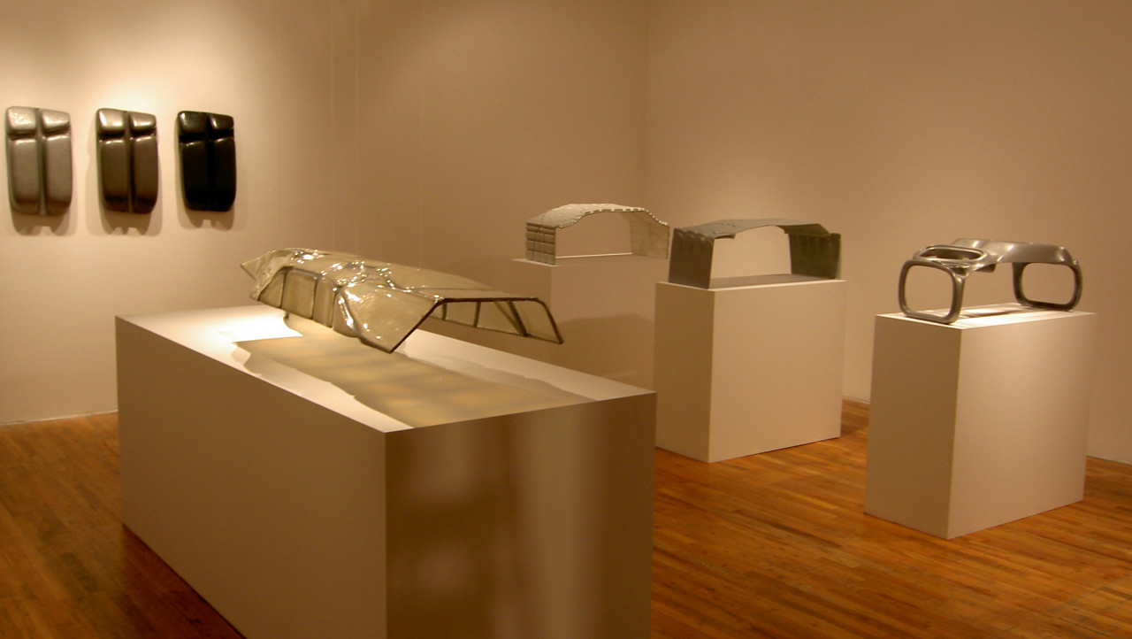Four abstract sculptures constructed from neutral-toned materials are displayed atop white virtrines. Three more sculptures are hung in a row against a white wall.