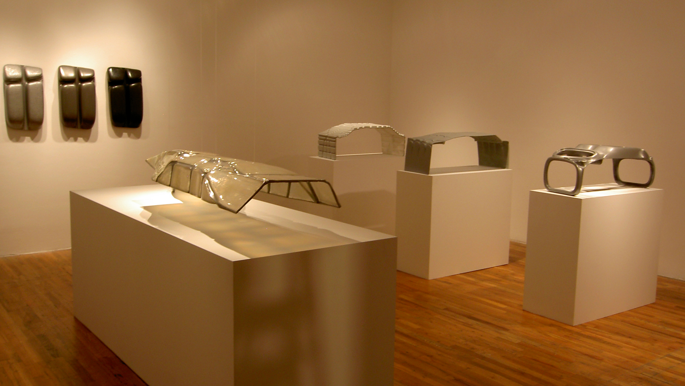 Four abstract sculptures constructed from neutral-toned materials are displayed atop white virtrines. Three more sculptures are hung in a row against a white wall.