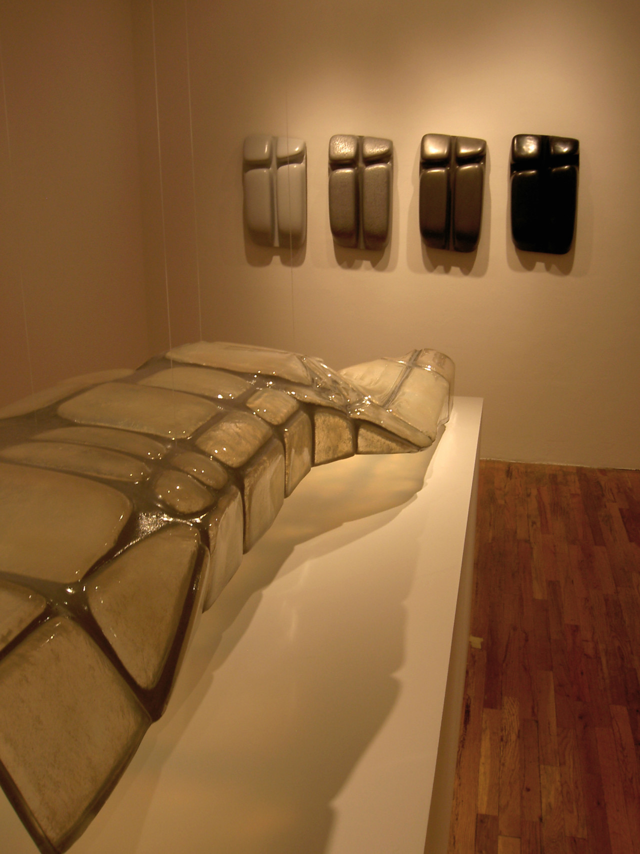 In the foreground, a clear and grey sculpture rests on top of a white pedestal. In the background, four additional sculptures in arranged from light to dark tones, hang in a row.