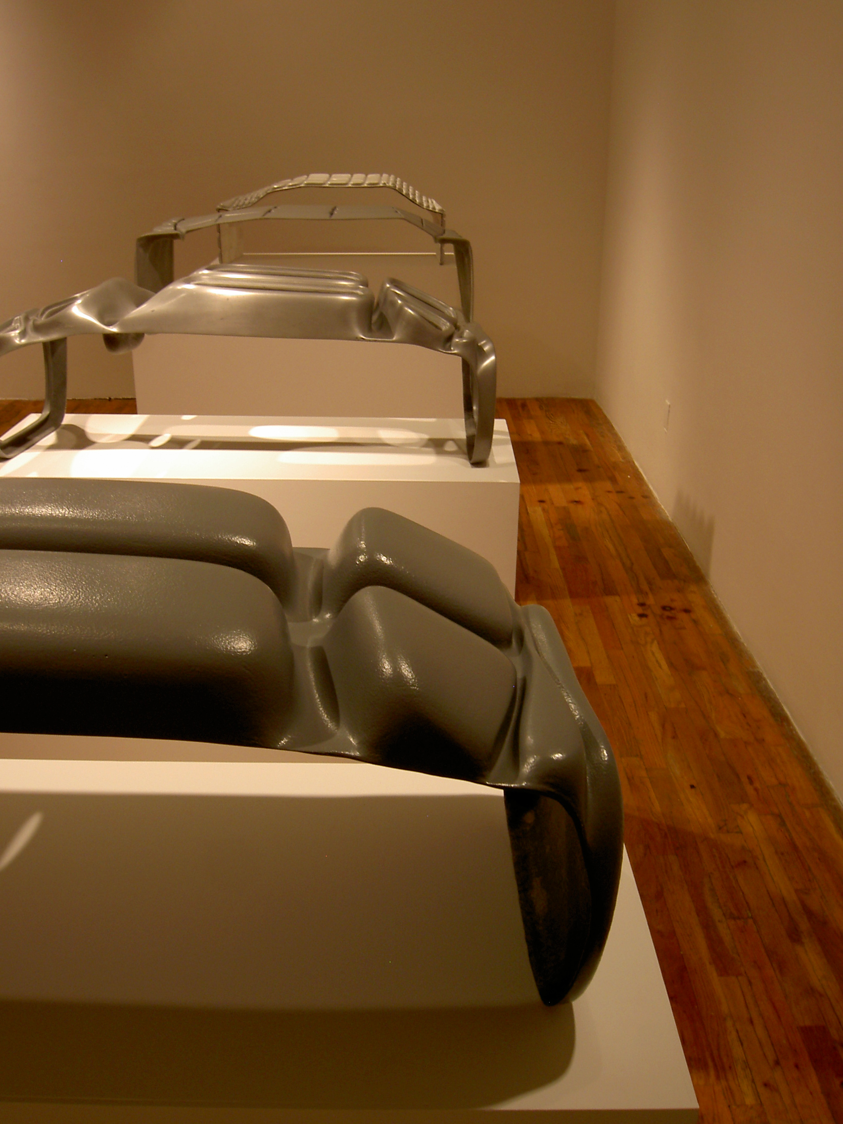 Four sculptures, each roughly the same size and configuration, are displayed on top of white vitrines. Each sculpture is constructed from a different material, with sy