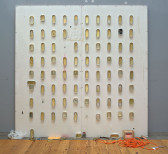 A large white MDF board rests against a gray wall. A series of metal sardine cans are placed across the MDF board in a loose grid. 
