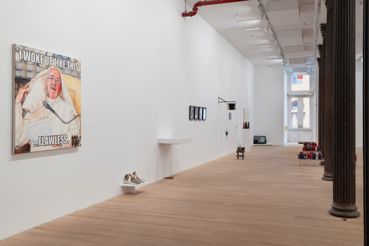 The gallery space displays an assortment of artworks on the left wall. On the far left, closest to the viewer, is a painting of Pope Francis with a top text reading "I WOKE UP LIKE THIS" and a bottom text reading "...FLAWLESS." An assortment of art objects, shelves, framed paintings, sculptural installations, and digital media recede into the gallery space along this wall. On the right a row of columns recede into the gallery space.