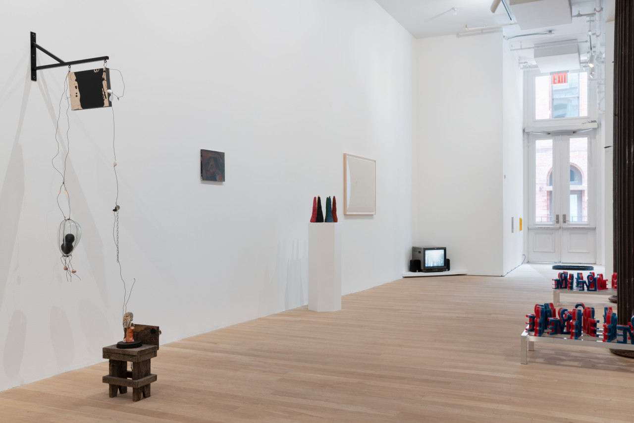 Along the gallery wall is an assemblage of objects hanging off the wall and to the floor connected by metal thread. Further down the wall is a painting, a plinth of colorful sculptures, a framed artwork, and a television playing a video in the corner. In the back is a glimpse of an alcove with three artworks appearing as colorful stripes, grey and yellow on the wall, and black on the floor.