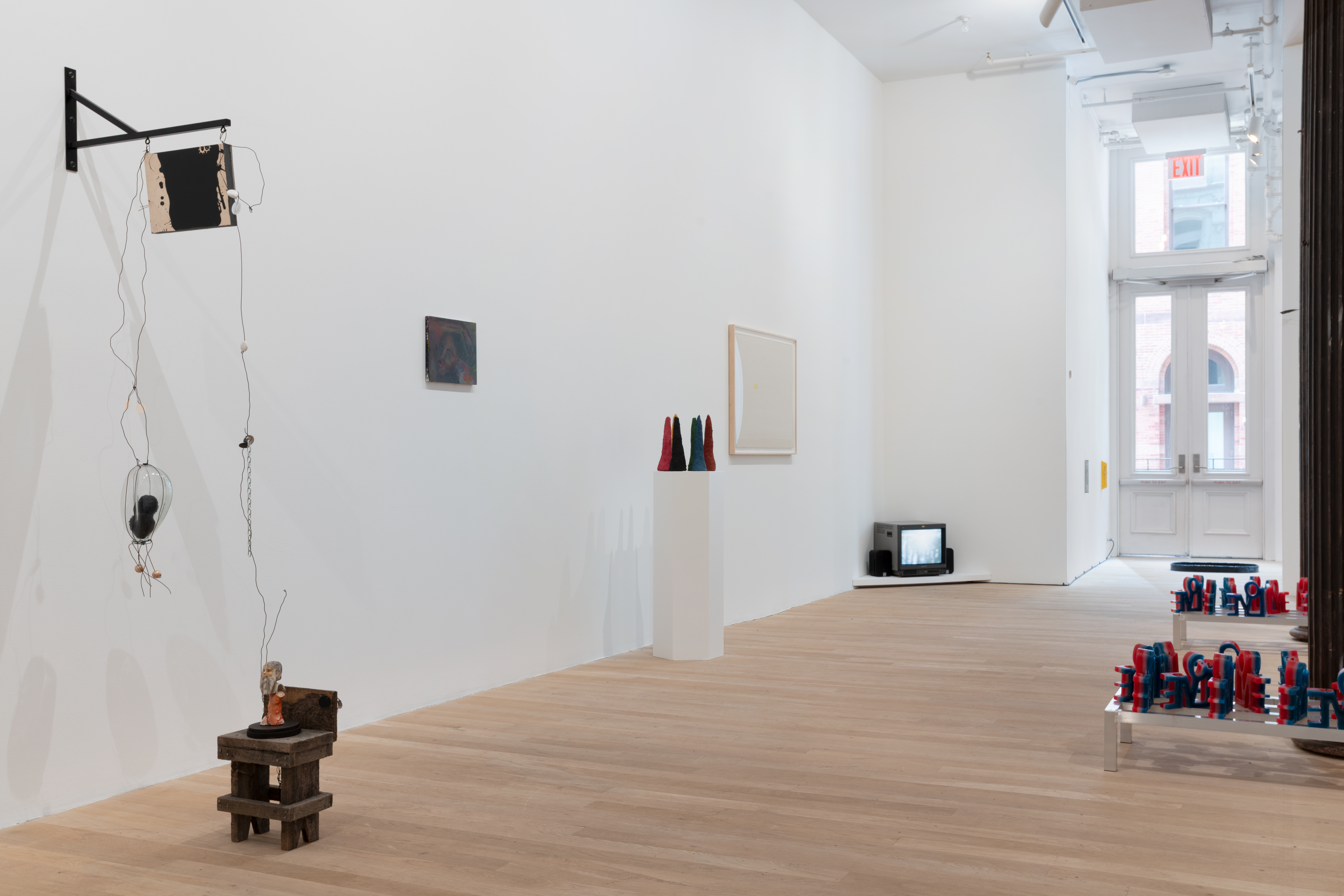 Along the gallery wall is an assemblage of objects hanging off the wall and to the floor connected by metal thread. Further down the wall is a painting, a plinth of colorful sculptures, a framed artwork, and a television playing a video in the corner. In the back is a glimpse of an alcove with three artworks appearing as colorful stripes, grey and yellow on the wall, and black on the floor. 