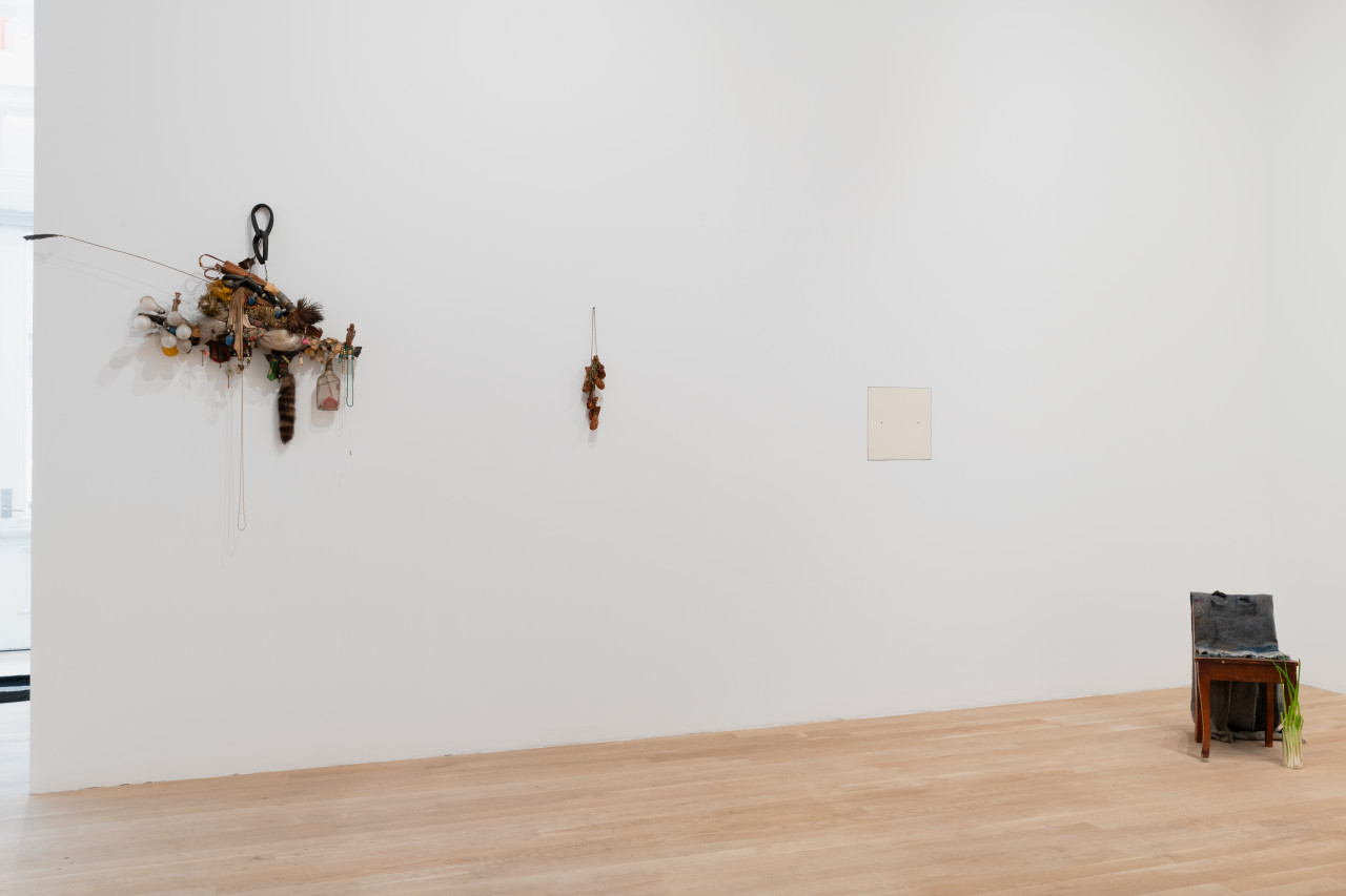 Along the gallery wall from left to right is an assemblage scultpure of found objects ranging from lightbulbs to racoon tails. To the right is a sculpture of chicken wings dangling from the wall. Further along is an offwhite square nearly flush with the wall. And on the floor sits a chair adorned with cloth and green onion.