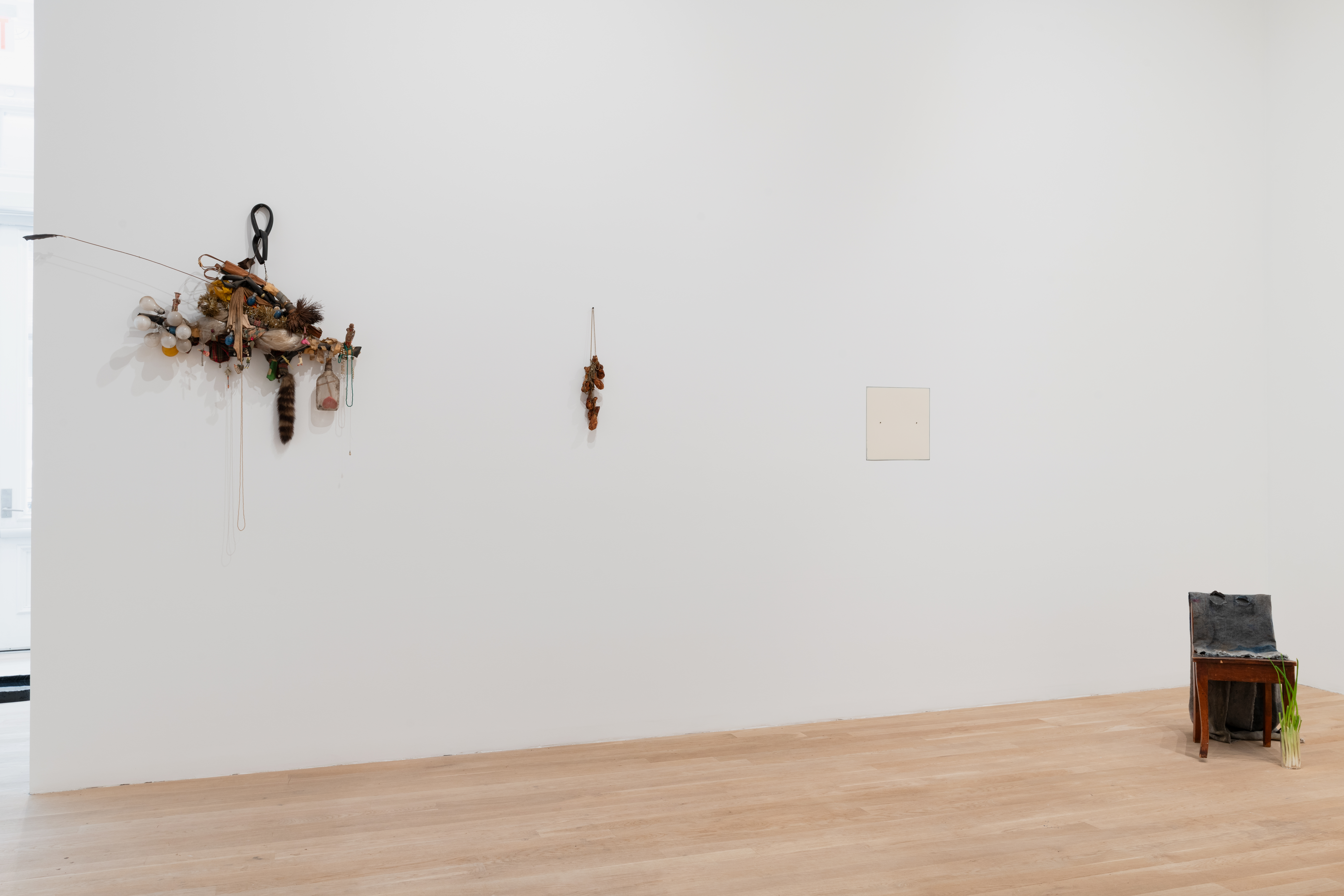 Along the gallery wall from left to right is an assemblage scultpure of found objects ranging from lightbulbs to racoon tails. To the right is a sculpture of chicken wings dangling from the wall. Further along is an offwhite square nearly flush with the wall. And on the floor sits a chair adorned with cloth and green onion.