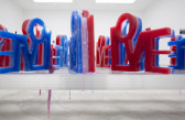 A series of candles, each made of layers of red, white, and blue wax, are placed on an aluminum shelf. From the front, the shape of the candles spell out 