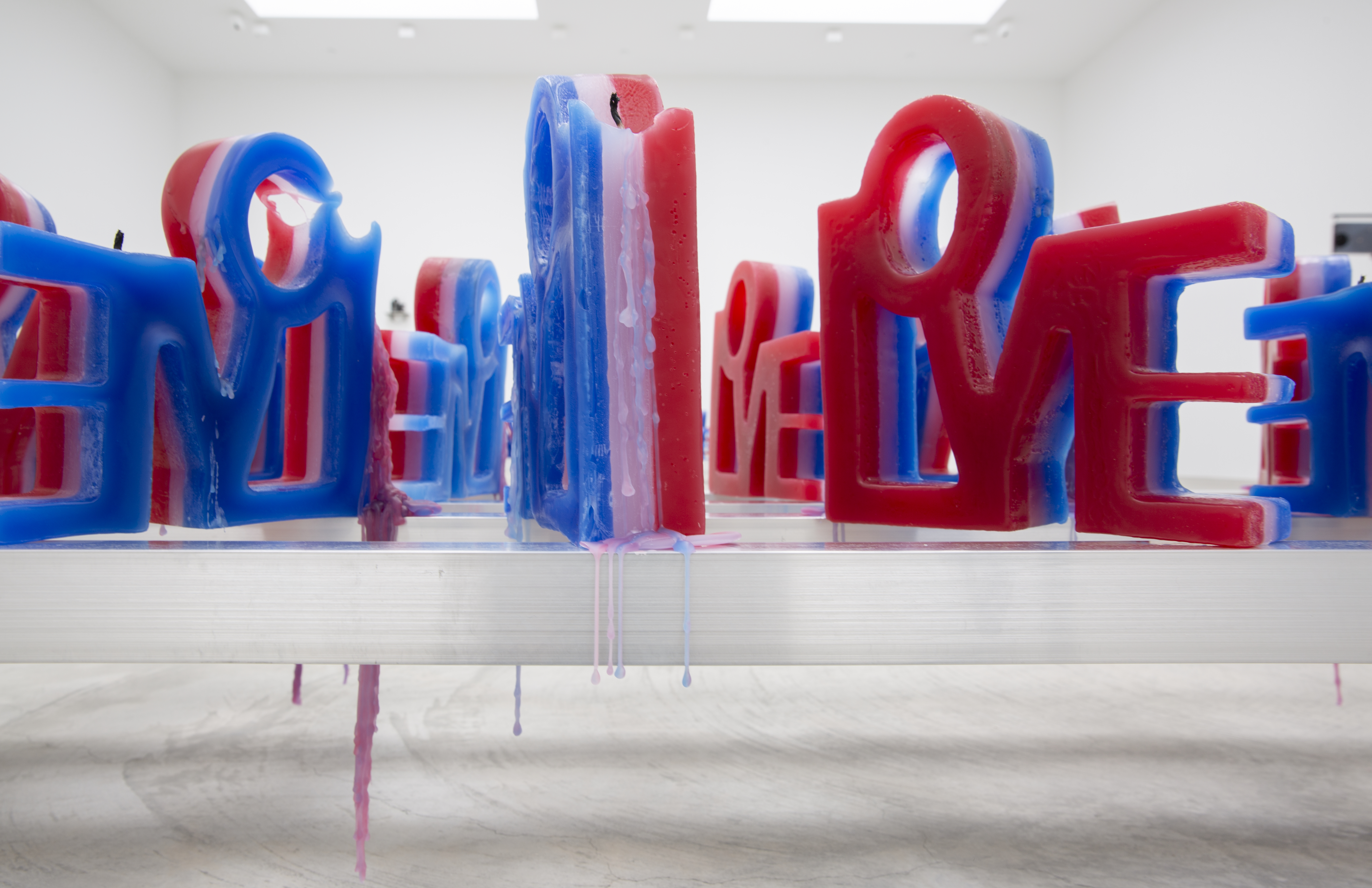 A series of candles, each made of layers of red, white, and blue wax, are placed on an aluminum shelf. From the front, the shape of the candles spell out 