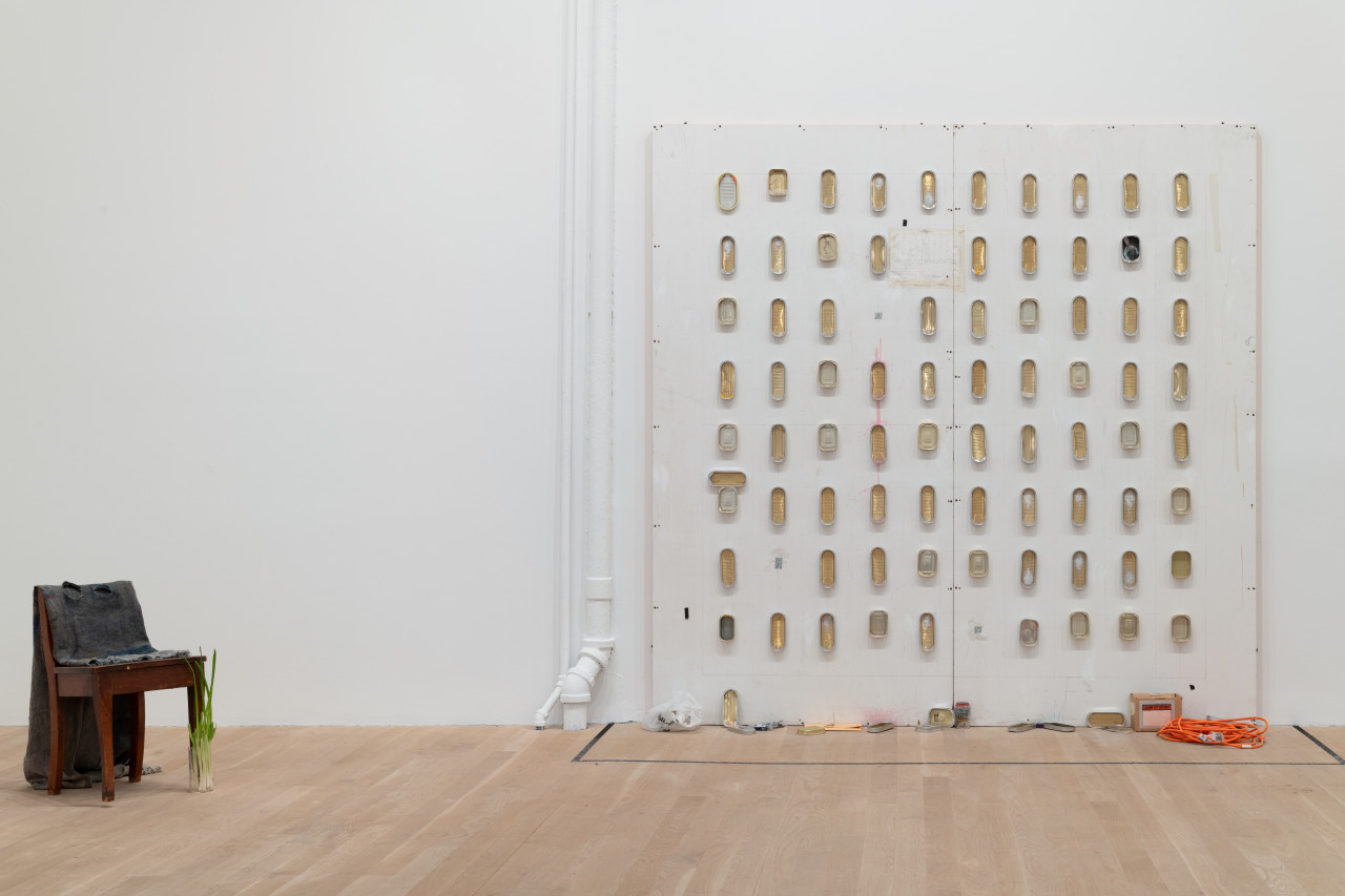 To the left sits a chair adorned in cloth and green onion. On the gallery wall is a square MDF wall with graddied sardine cans along in. On the floor in front of this MDF wall is an assortment of objects: bag, box, electrical cord, and more sardine cans.