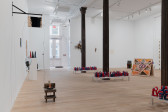 An assortment of artworks are seen in the large gallery space. On the left, an assemblage of materials affixed to the wall on floor connected through a metal thread. Behind are framed works and a plinth displaying colorful sculptures. In the right of the gallery are red, white, and blue wax candles spelling 