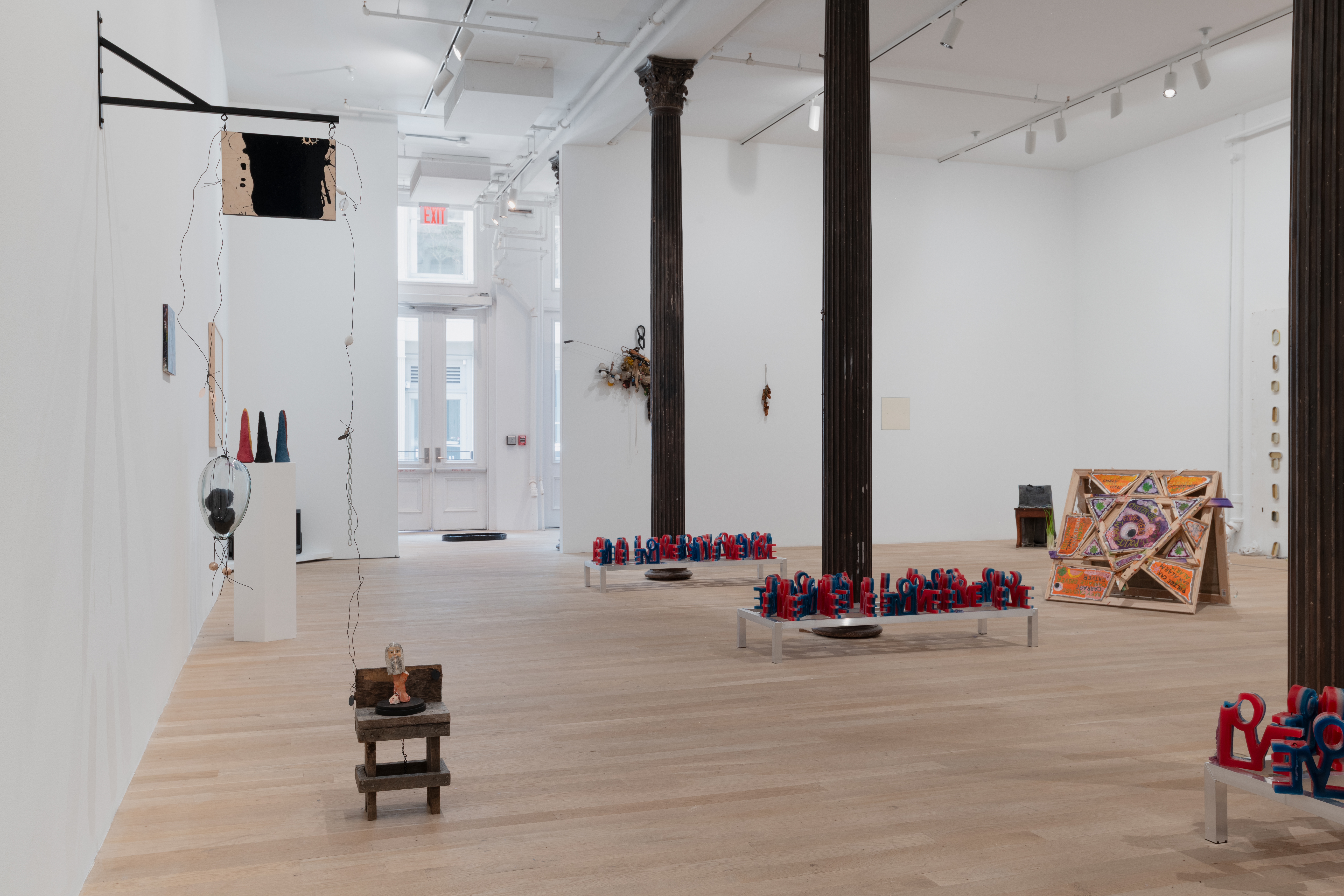 An assortment of artworks are seen in the large gallery space. On the left, an assemblage of materials affixed to the wall on floor connected through a metal thread. Behind are framed works and a plinth displaying colorful sculptures. In the right of the gallery are red, white, and blue wax candles spelling 