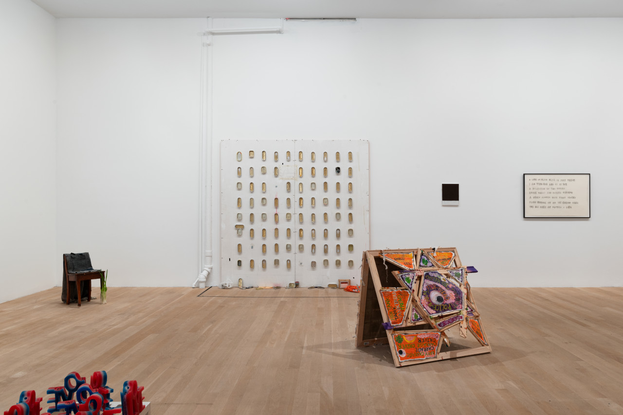 In the center of the gallery space wall rests a large, square MDF wall with a grid of empty sardine cans. To the left of this MDF wall is a chair adorned with various objects, and in the forefront red, white, and blue wax candles spelling "LOVE." To the right of the MDF wall is an artwork on the floor in the spce of the Star of David with painted text on top. On the wall is a black and white painting and a framed text drawing.