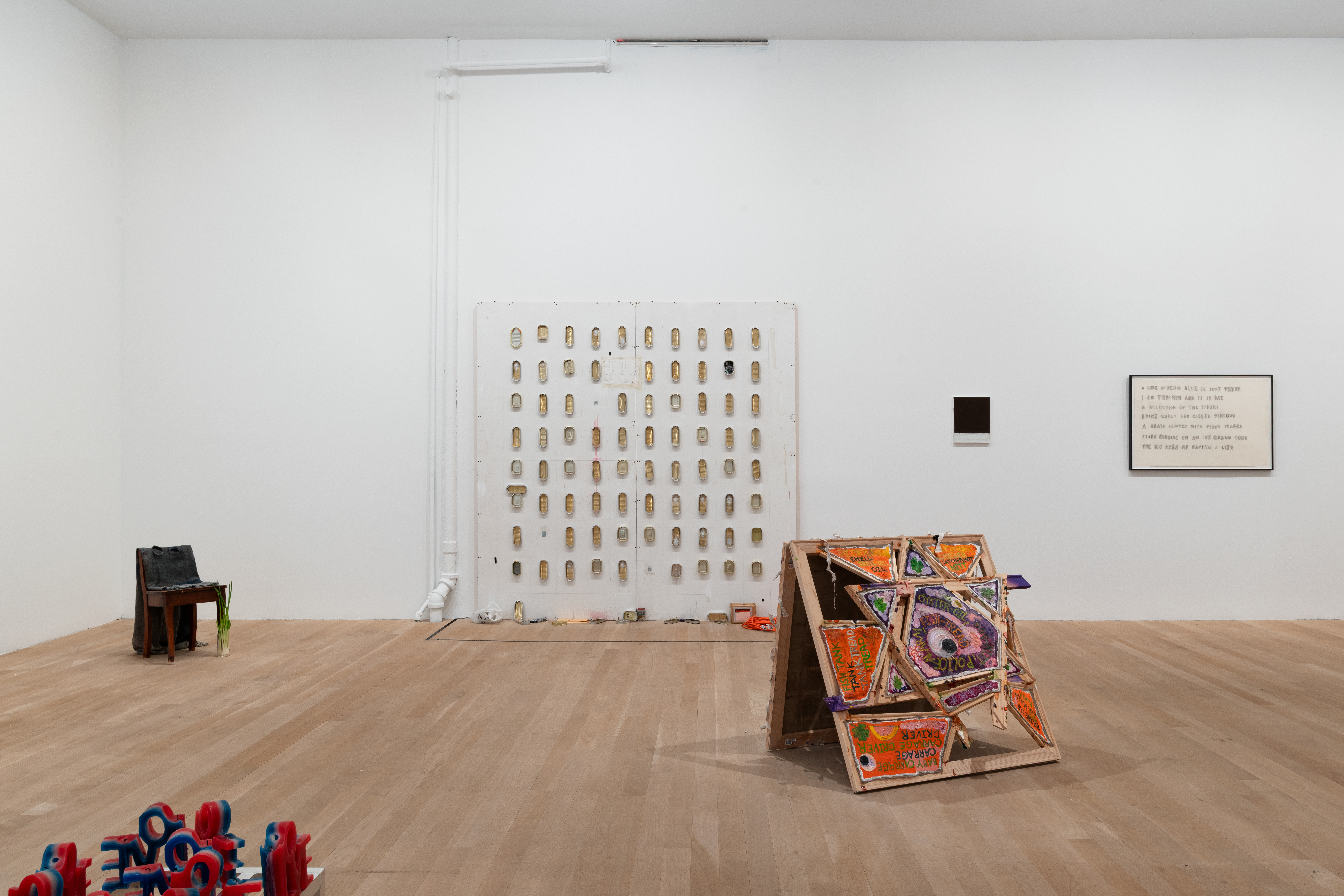 In the center of the gallery space wall rests a large, square MDF wall with a grid of empty sardine cans. To the left of this MDF wall is a chair adorned with various objects, and in the forefront red, white, and blue wax candles spelling 