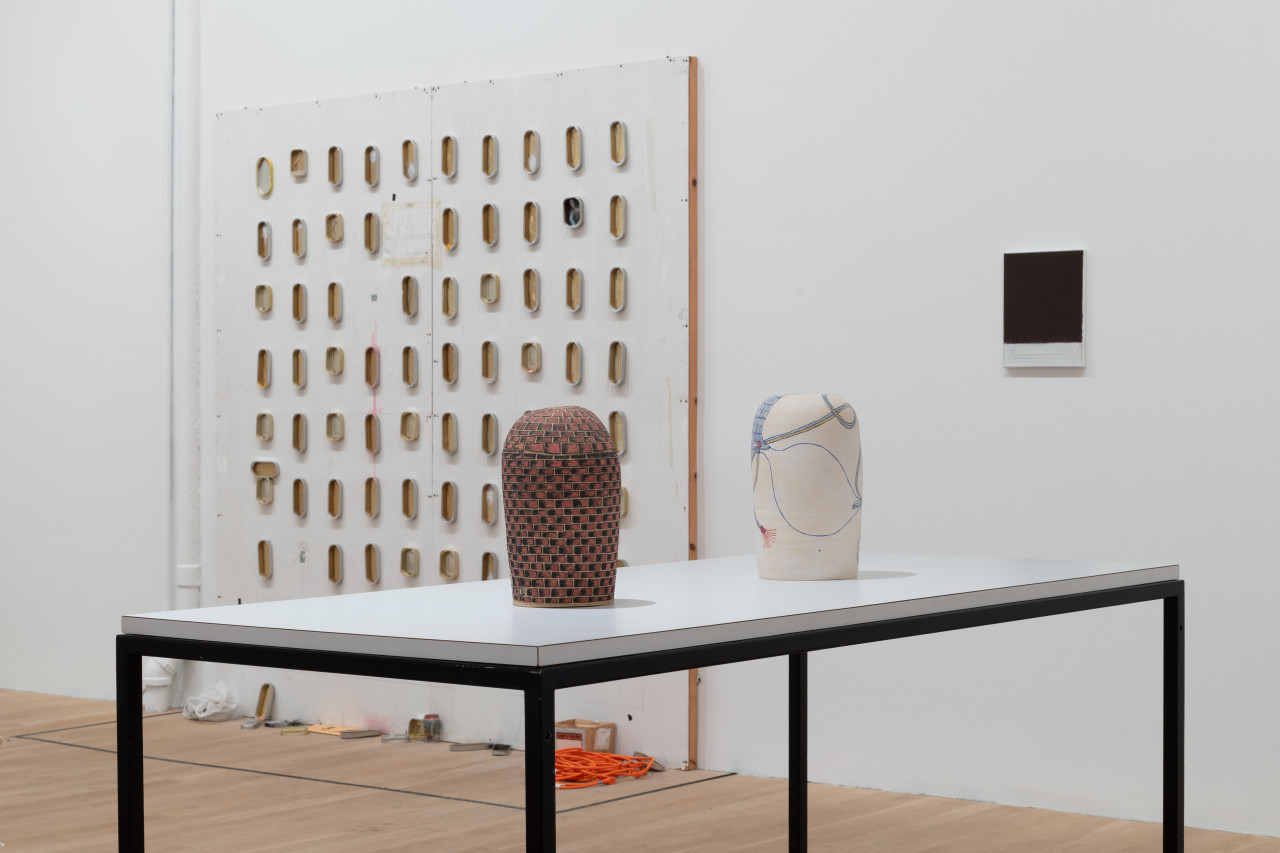 Two ceramic works sit in the foreground on a table. The left ceramic is painted with bricks and the right ceramic is painted with line. In the back is a square, MDF wall of gridded sardine cans and a black and white painting.