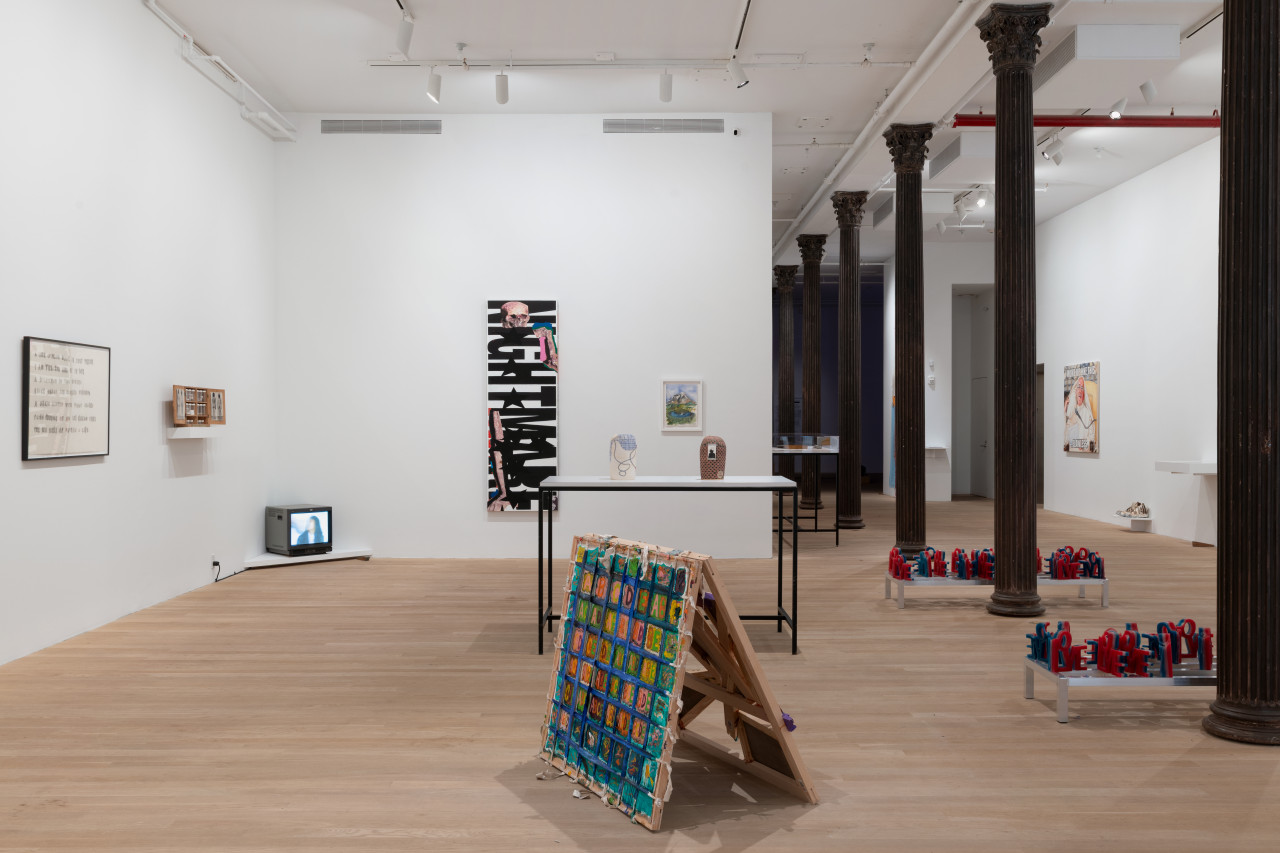 An assortment of artworks are seen in the large gallery space. On the left wall rests a framed text drawing, a shelf holding a display of photographs, and in the corner a television playing a video. The wall to the right displays two paintings. On the floor in front of these walls in a table with two ceramic works and gridded artwork on the floor. The space is divided by columns receding into the back of the gallery space.