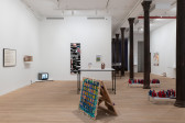 An assortment of artworks are seen in the large gallery space. On the left wall rests a framed text drawing, a shelf holding a display of photographs, and in the corner a television playing a video. The wall to the right displays two paintings. On the floor in front of these walls in a table with two ceramic works and gridded artwork on the floor. The space is divided by columns receding into the back of the gallery space.