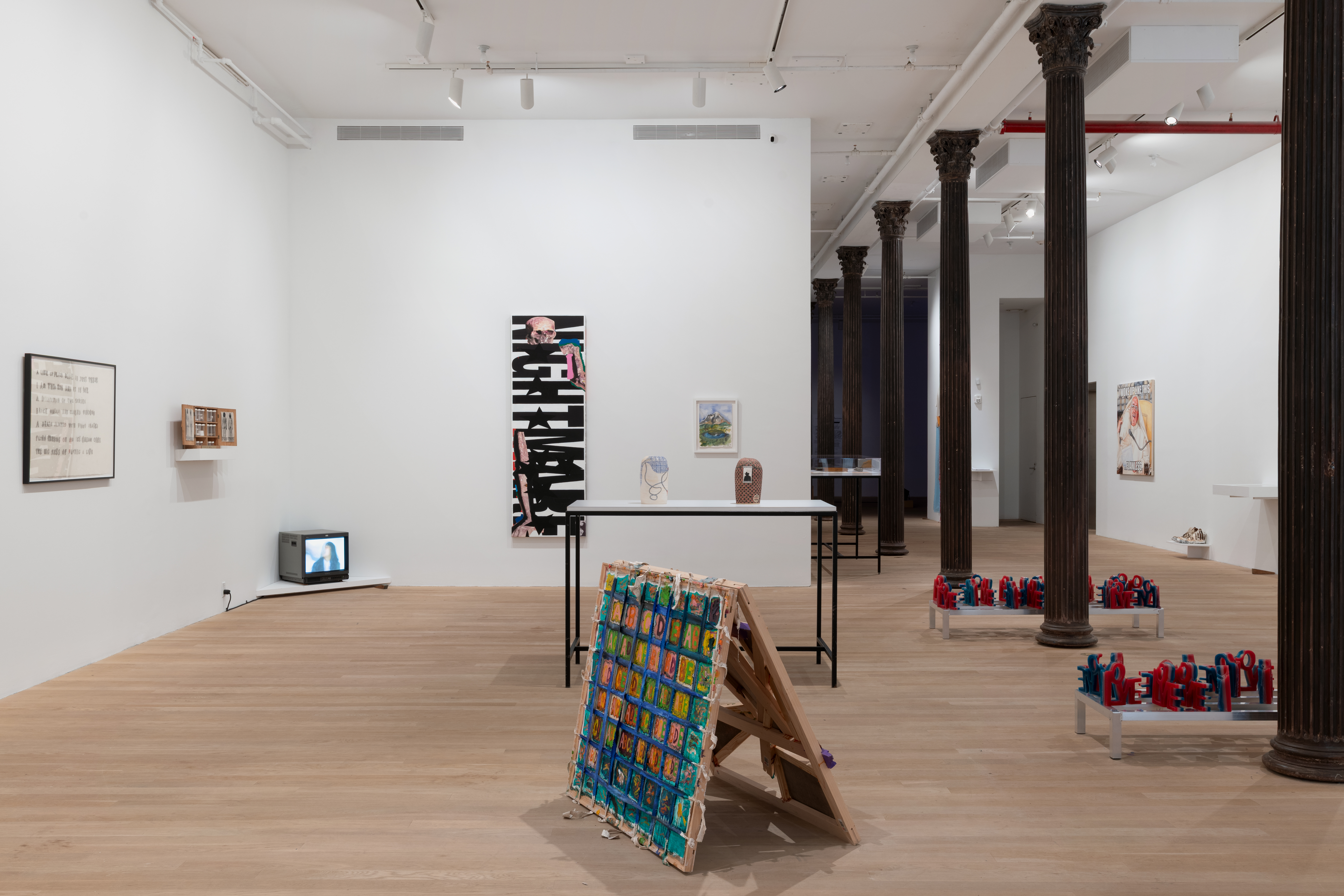 An assortment of artworks are seen in the large gallery space. On the left wall rests a framed text drawing, a shelf holding a display of photographs, and in the corner a television playing a video. The wall to the right displays two paintings. On the floor in front of these walls in a table with two ceramic works and gridded artwork on the floor. The space is divided by columns receding into the back of the gallery space.