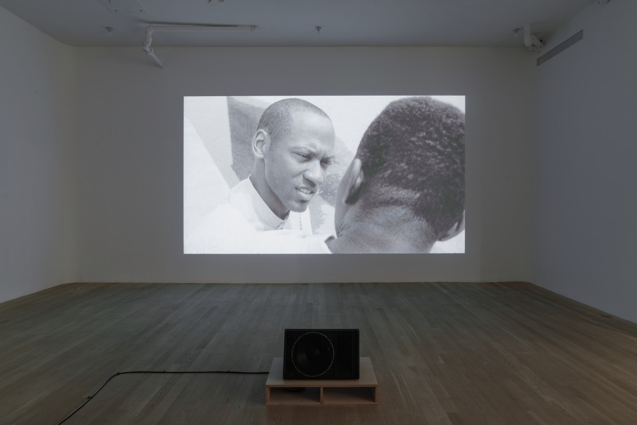 A projected video displays a black and white still of two black men looking at each other. We see the back of one of these men