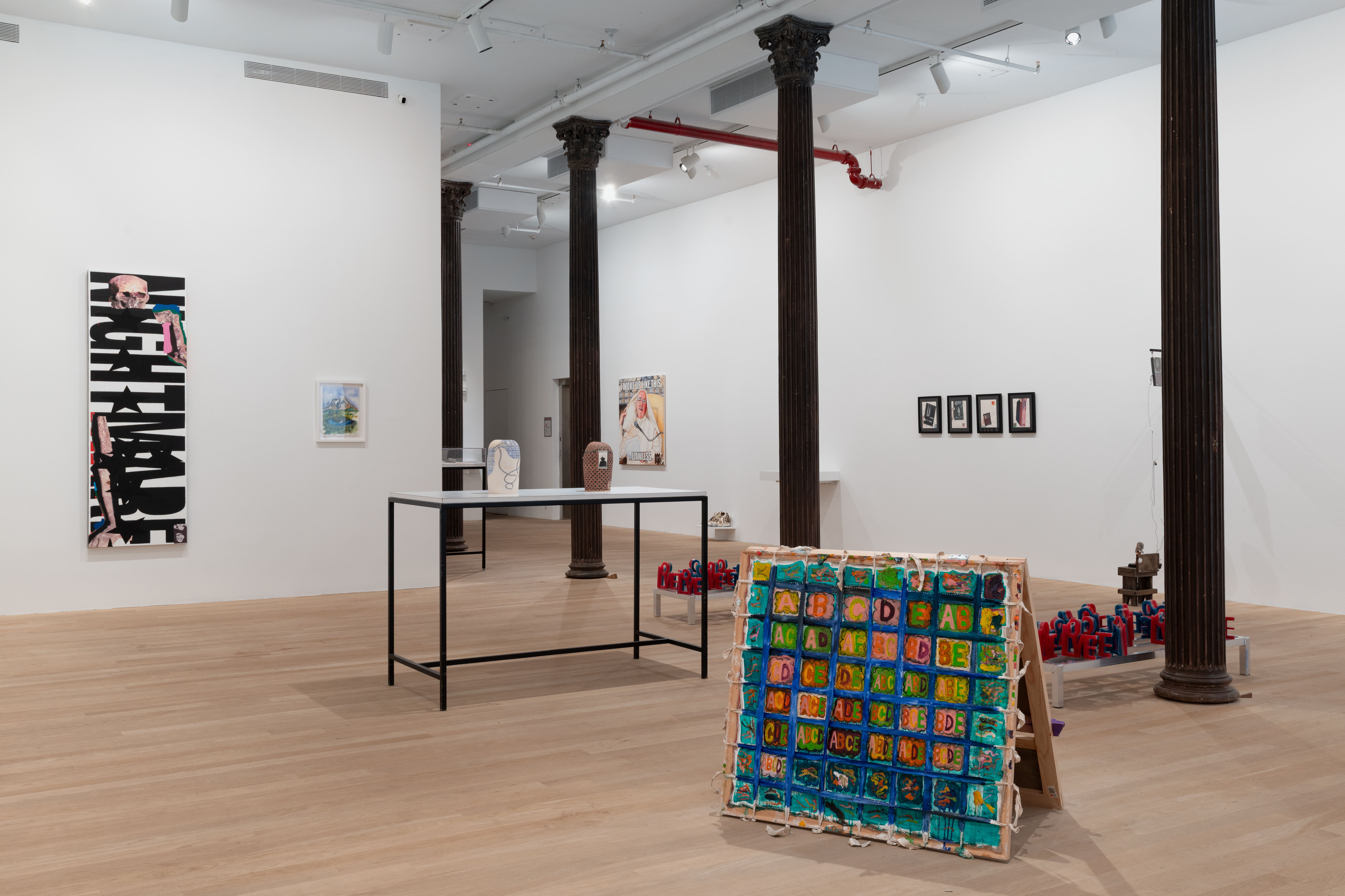 An assortment of artworks are seen in the large gallery space. On the left wall, a painting reading 