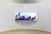 A lightbox displaying a group of sitting and standing individuals. Behind them is a wall of small photographs and artworks.