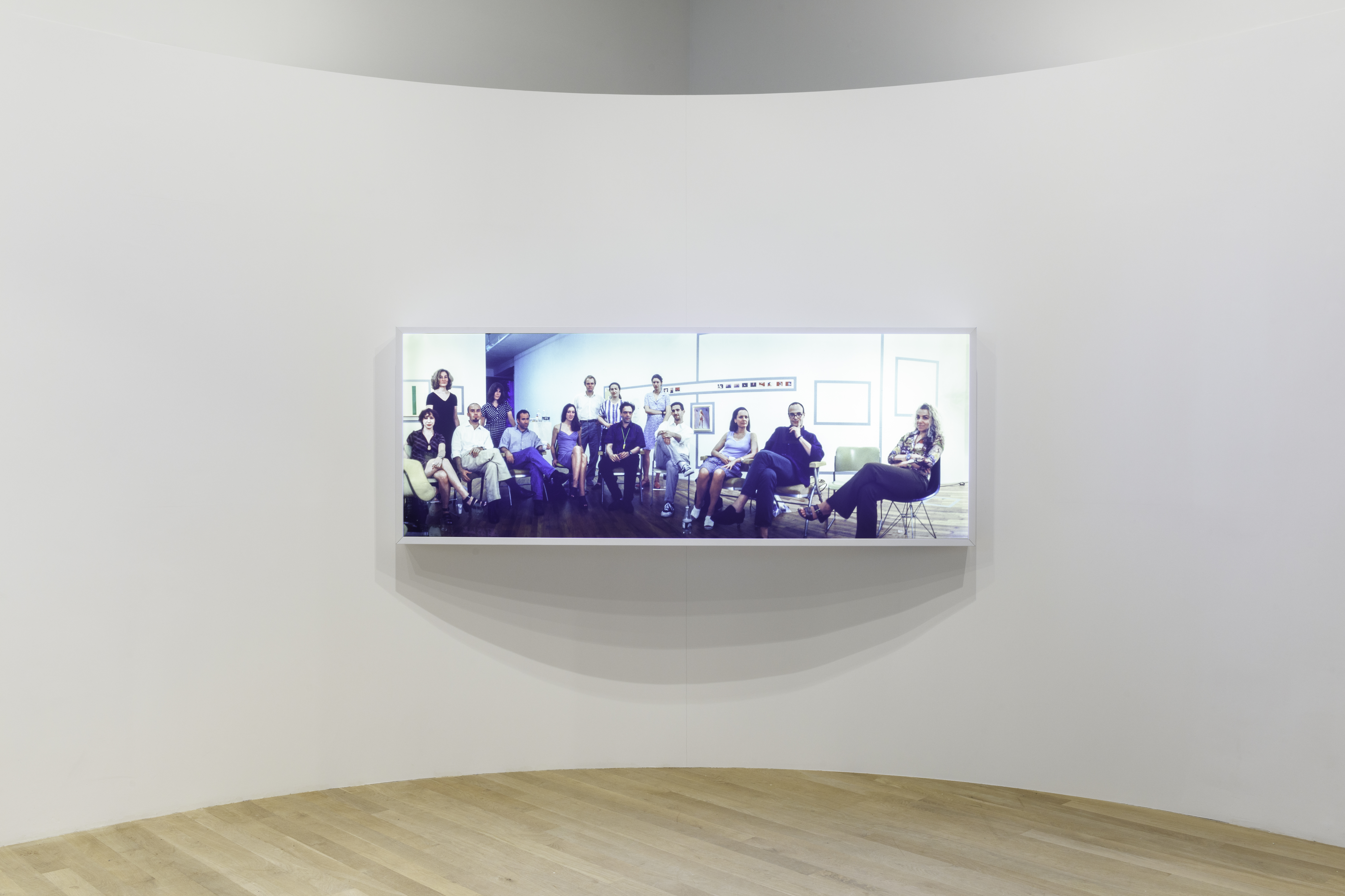 A lightbox displaying a group of sitting and standing individuals. Behind them is a wall of small photographs and artworks.