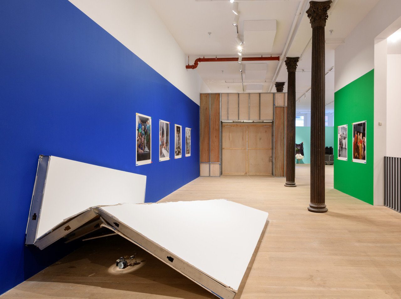 A view of a small drum set lit by a spotlight underneath a demolished wall, blue and green gallery walls displaying dynamic images of individuals hung by large push pins.