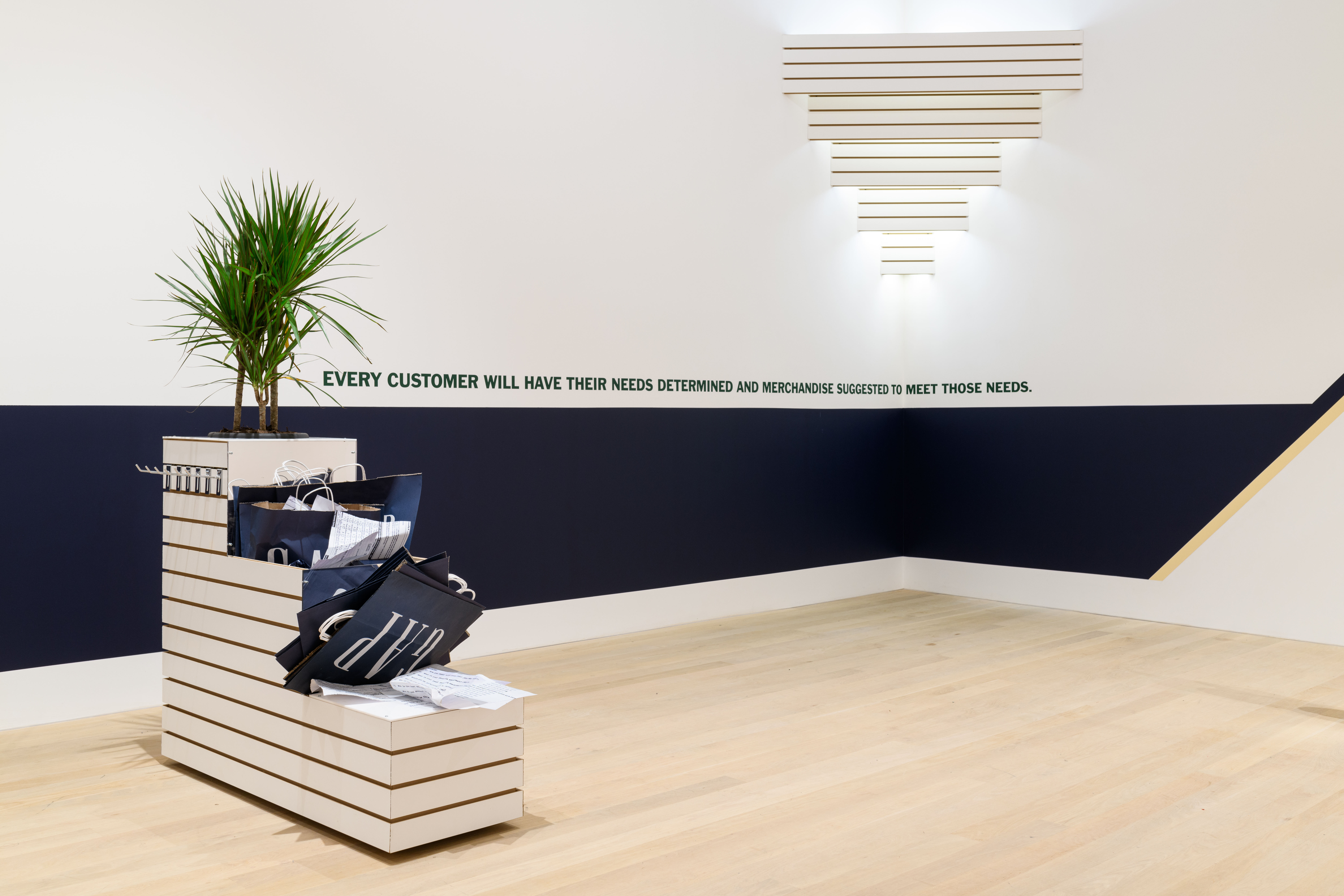 A small store display shelf stuffed with Gap clothing store bags and a small palm tree. On the wall, a blue band wraps around to the adjacent wall with text that reads 