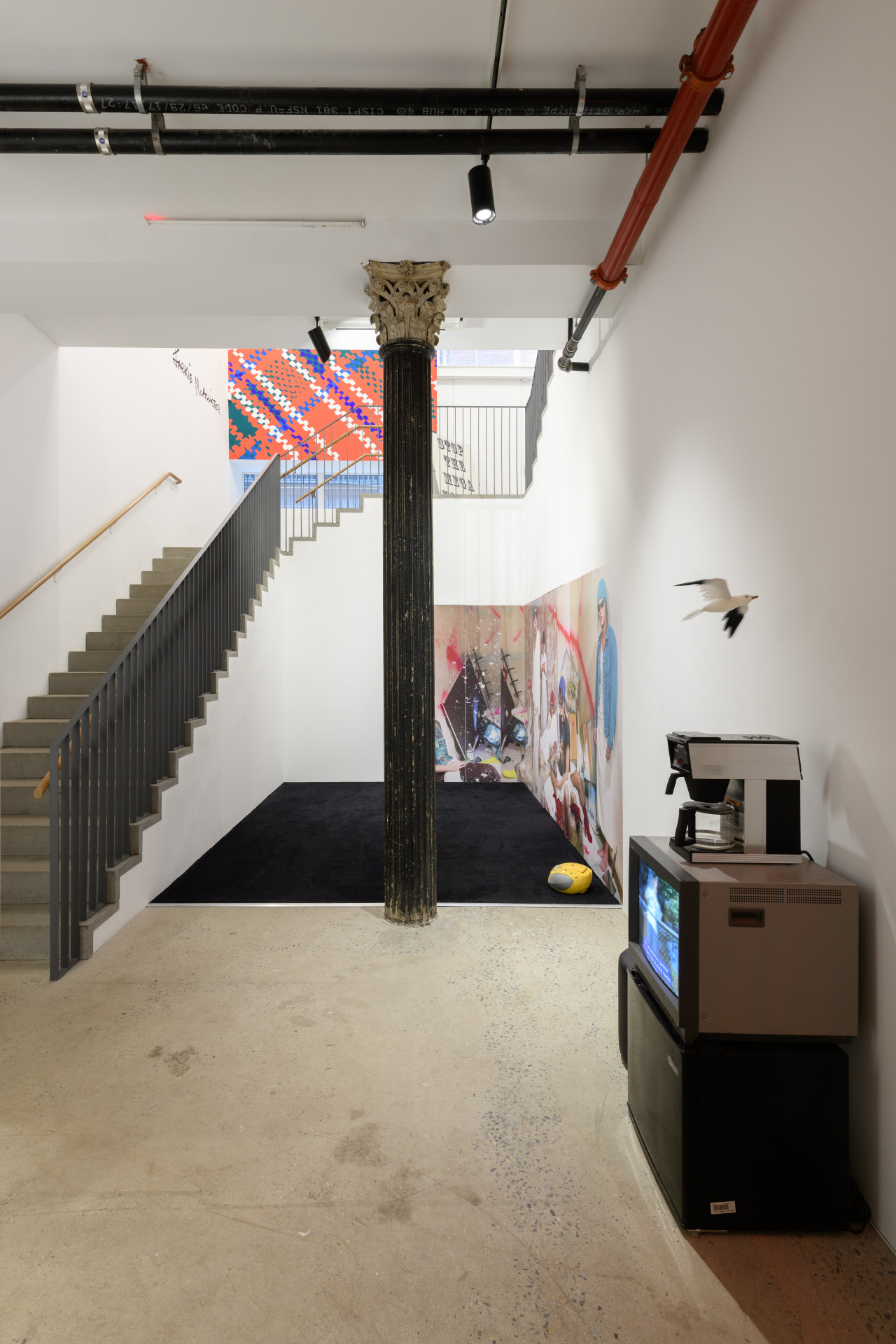 A view of multiple works displayed in a multilevel gallery space. An old tv monitor, a coffee machine, and an artificial seagull are placed near a yellow boombox sitting on a black rug. Beside the boombox is a large image of a group of individuals in a defaced bedroom.