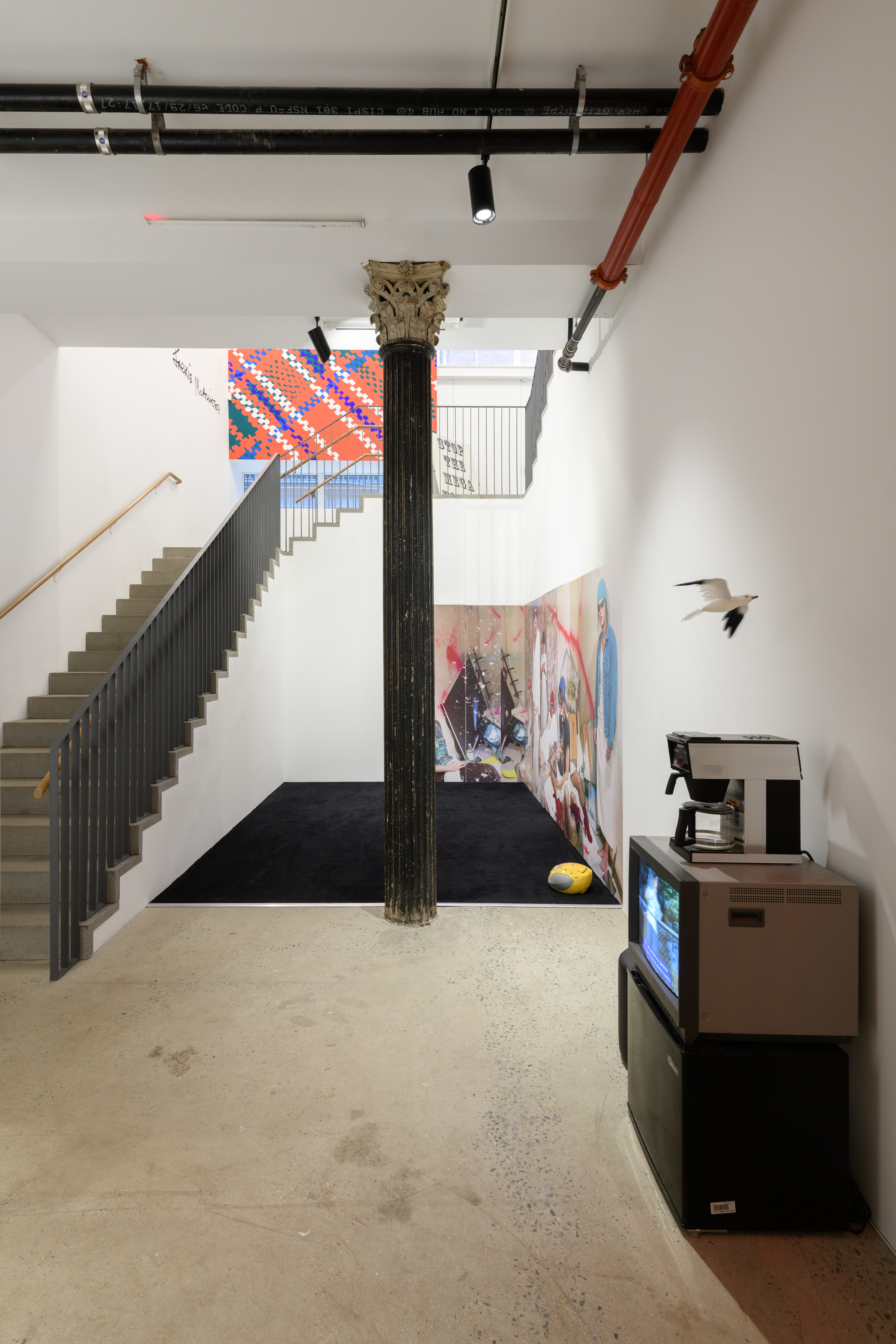 A view of multiple works displayed in a multilevel gallery space. An old tv monitor, a coffee machine, and an artificial seagull are placed near a yellow boombox sitting on a black rug. Beside the boombox is a large image of a group of individuals in a defaced bedroom.