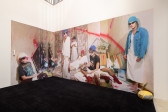 A view of a large image of multiple individuals sitting and standing in a defaced bedroom. A yellow boombox sits on a black carpet.
