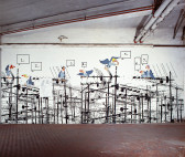 A painting on the wall of an industrial room depicts a network of television antennas, repeated horizontally. Cartoon birds sit atop the poles, each with a numbered speech bubble floating above their heads.