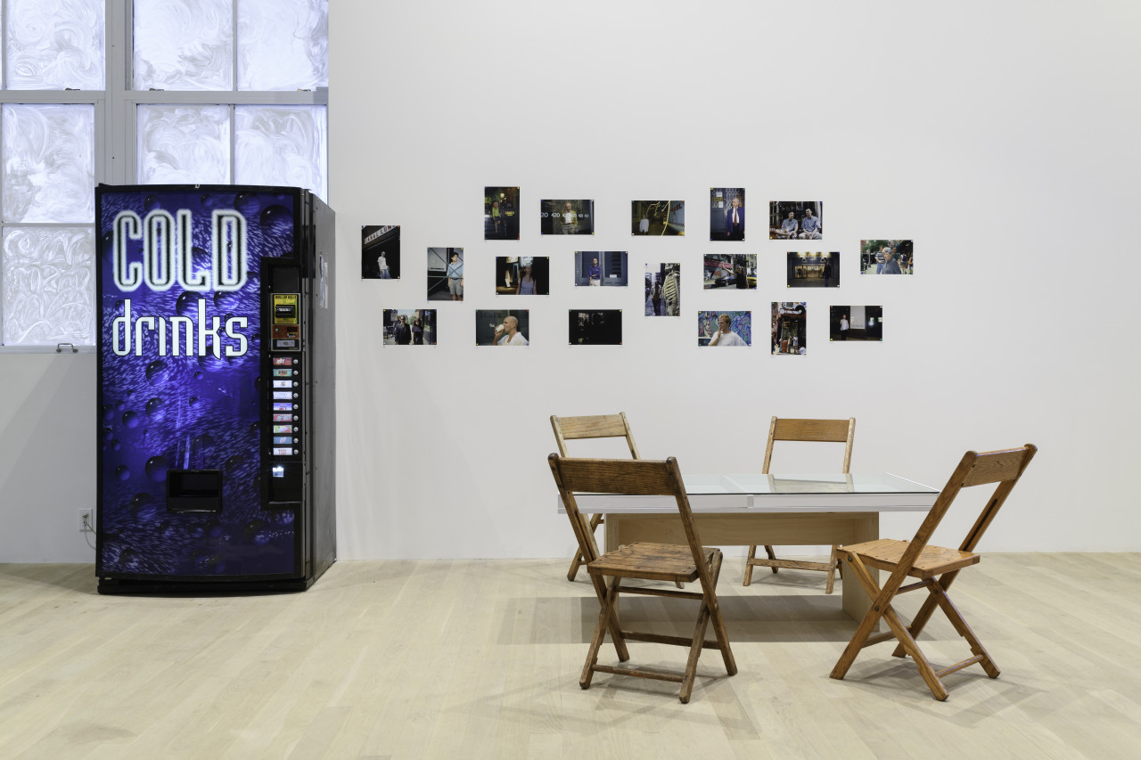 A view of a vending machine against a glazed over window, tiled images on a white gallery wall, and a glass table surrounded by four wooden chairs.