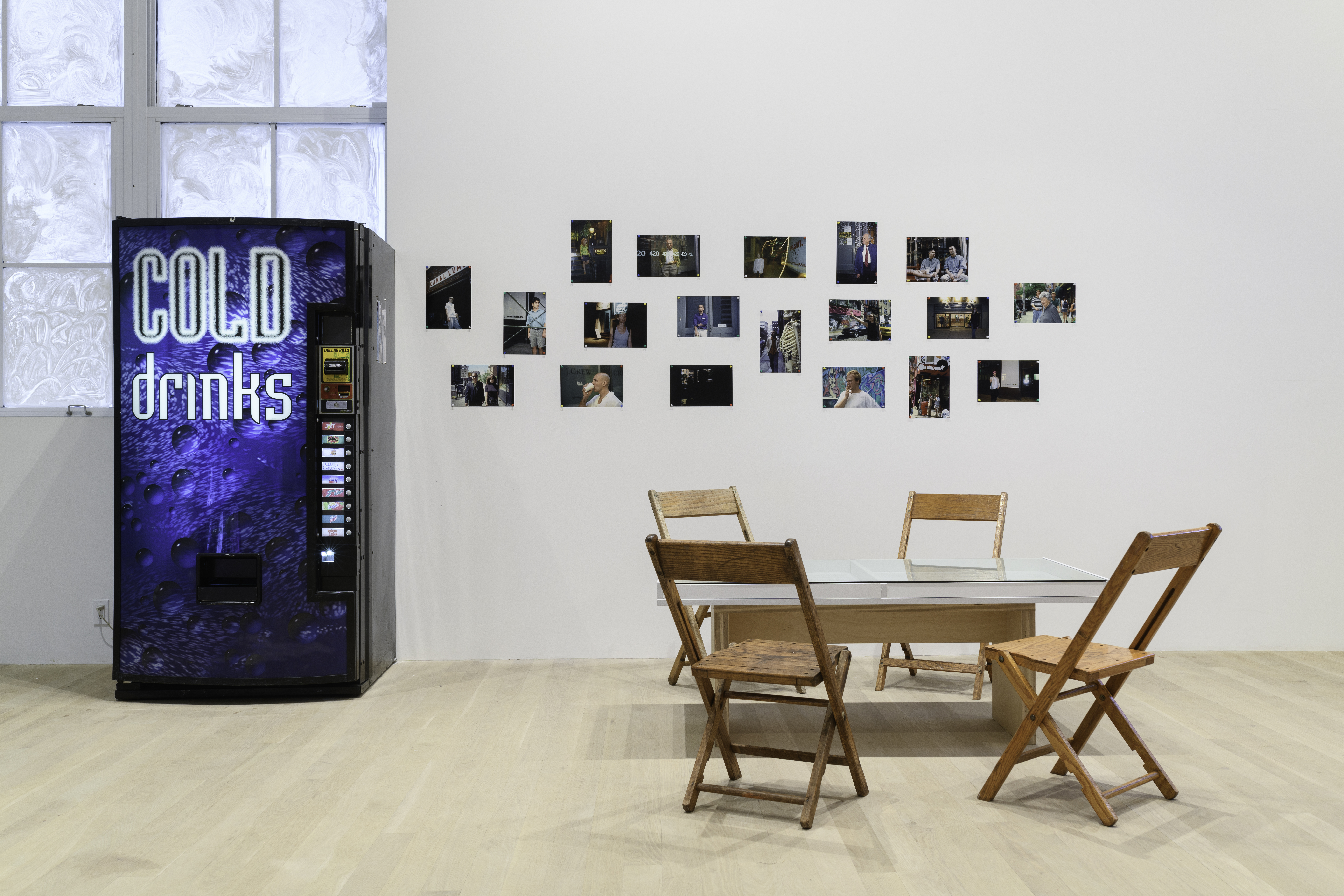 A view of a vending machine against a glazed over window, tiled images on a white gallery wall, and a glass table surrounded by four wooden chairs.