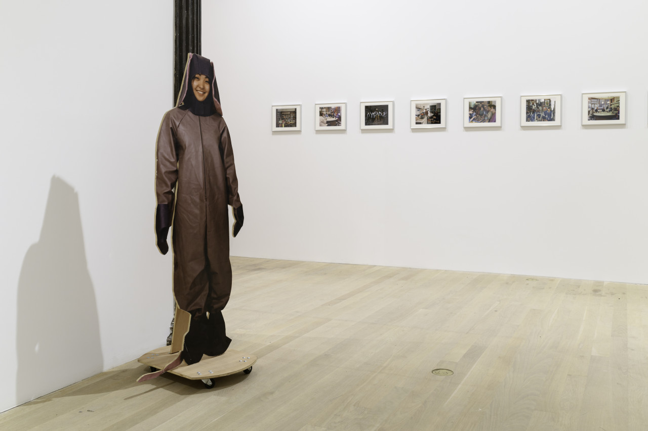 A view of a life like cutout of an individual dressed in an animal costume placed against a gallery column. Behind the cutout is a series of seven images displayed on a white gallery wall