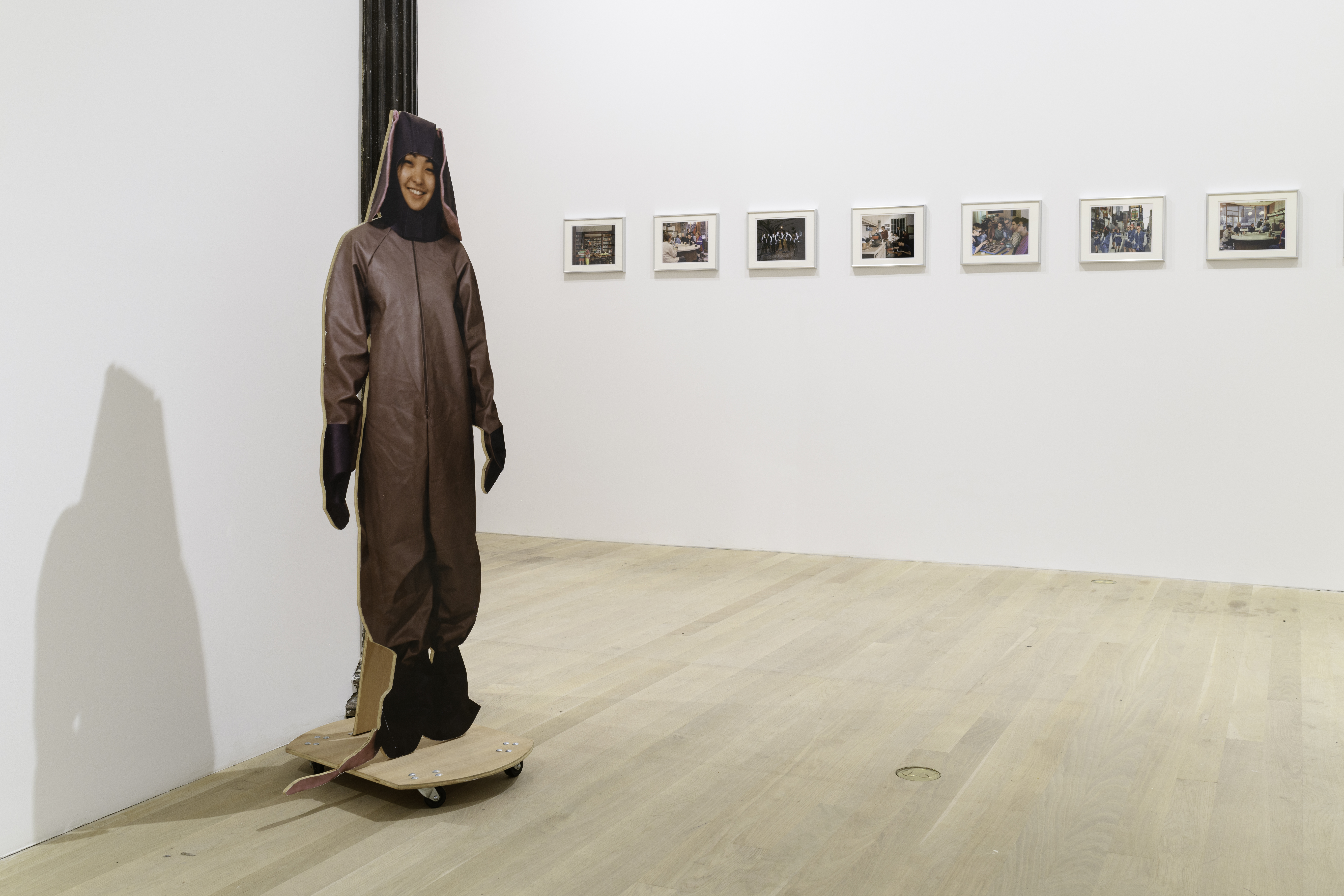 A view of a life like cutout of an individual dressed in an animal costume placed against a gallery column. Behind the cutout is a series of seven images displayed on a white gallery wall