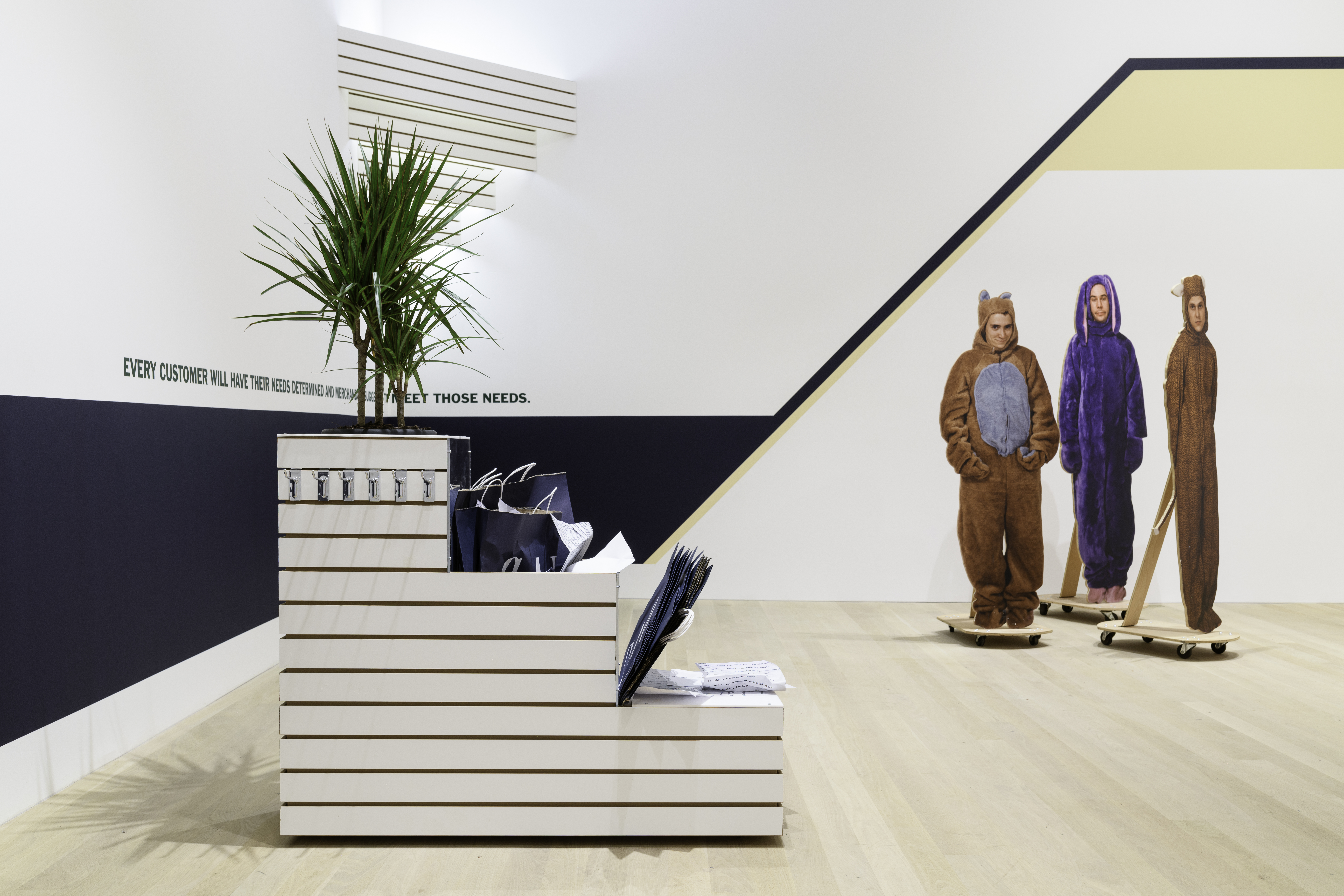 A view of a small store display shelf stuffed with Gap clothing store bags and a small palm tree. On the wall, a blue band wraps around to the adjacent wall with text that reads 