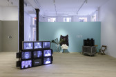 A view of multiple works including a black and white image on a beige wall, CRT monitors displaying individuals in a bluish hue, a tulip chair against a seafoam colored wall, and a single viewing station.