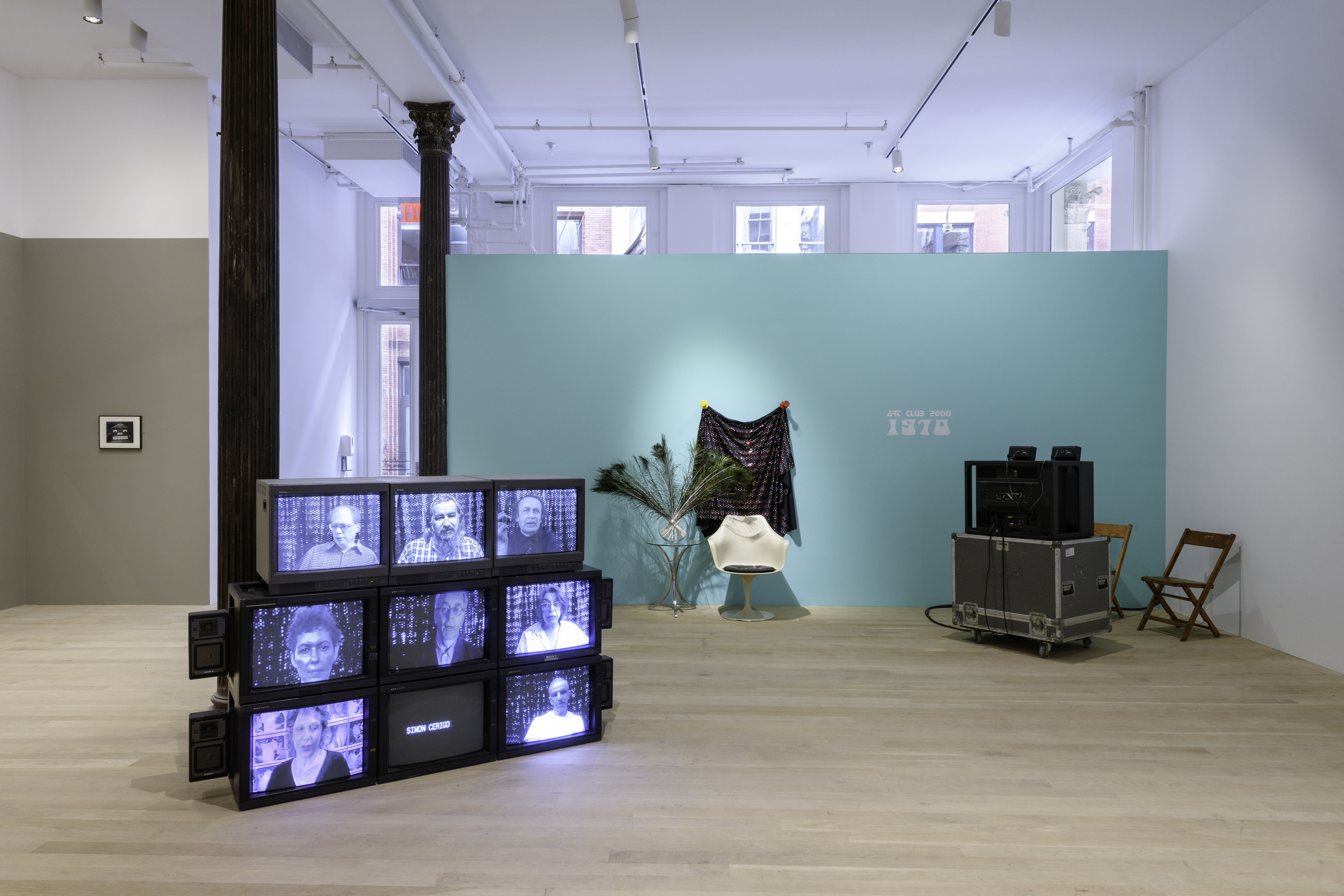 A view of multiple works including a black and white image on a beige wall, CRT monitors displaying individuals in a bluish hue, a tulip chair against a seafoam colored wall, and a single viewing station.