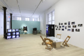 A view of multiple works including CRT monitors, a vending machine, a tulip chair against a seafoam colored wall, and a glass display table backed by tiled photographs.