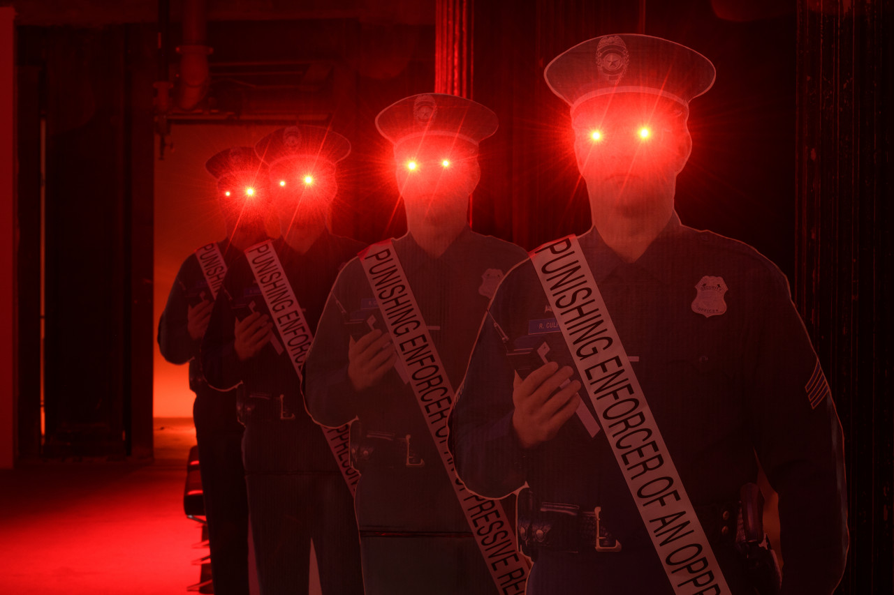 A view of four police officer cardboard cutouts with red eyes in a red lit room. They are wearing sashes that read "Punishing Enforcers of an Oppressive Regime."