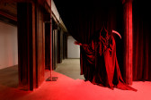 A grim reaper shrouded in deep red light pulls back a black curtain, beckoning one into a seperate vacant room.