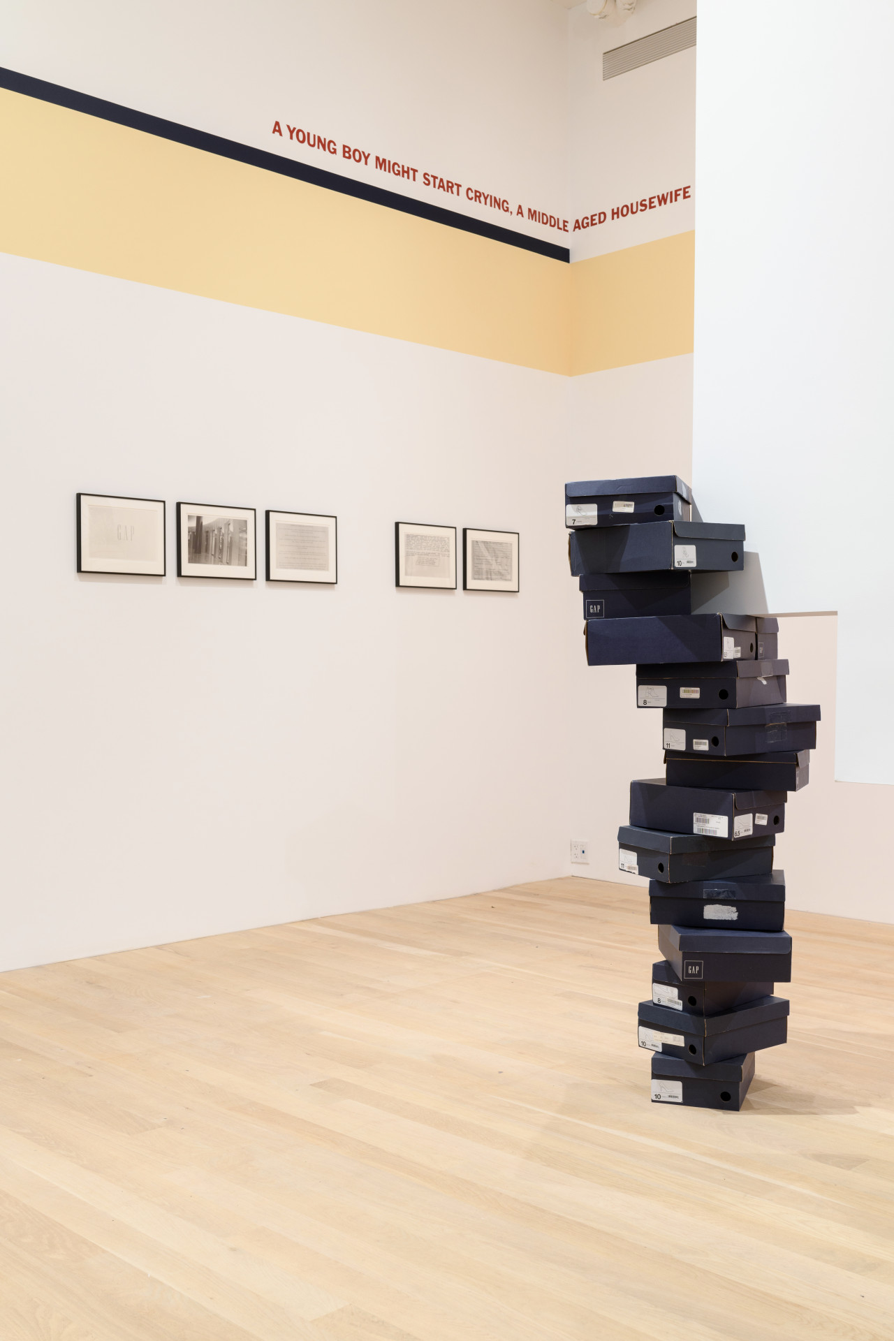 A display of Gap shoe boxes that meet an intersecting wall. Behind them, on the gallery wall is a series of images showing found documents. Above the images is a yellow band that wraps to the adjacent wall with text that reads "A youngboy might start crying, a middle aged housewife".