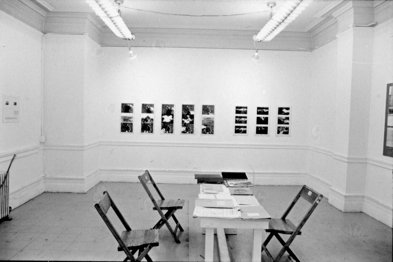 A photograph of a room hung with photographs and works on paper. In the center of the room is a table with folders and magazines and three chairs nearby.