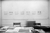 A photograph of a gallery wall hung with seven works on paper. In front of the wall are two chairs and a table with several envelopes and magazines.