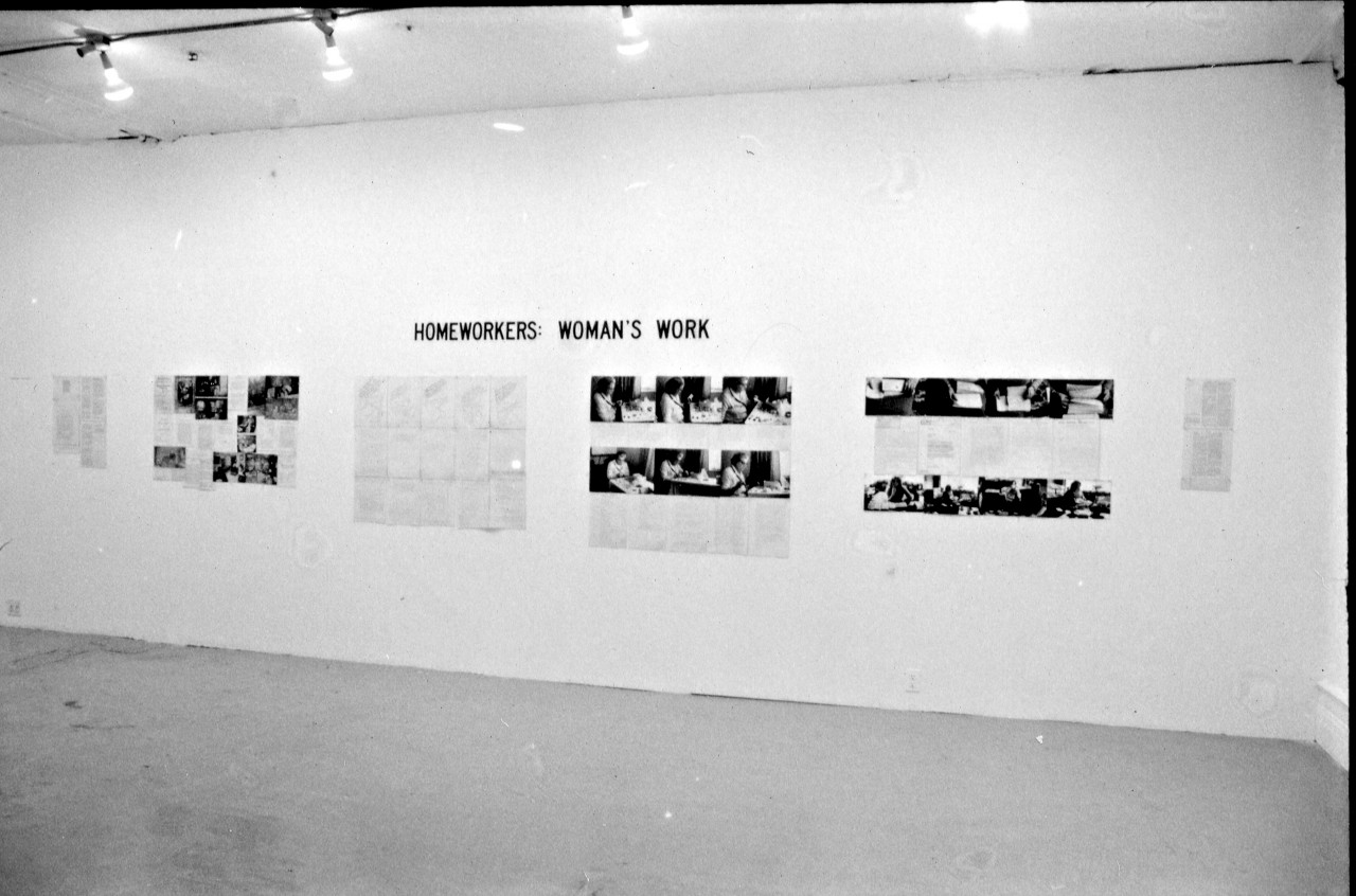 A photograph of a white gallery wall, hung with photographs and printed text. The title "HOMEWORKERS: WOMAN