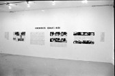 A photograph of a white gallery wall, hung with photographs and printed text. The title 