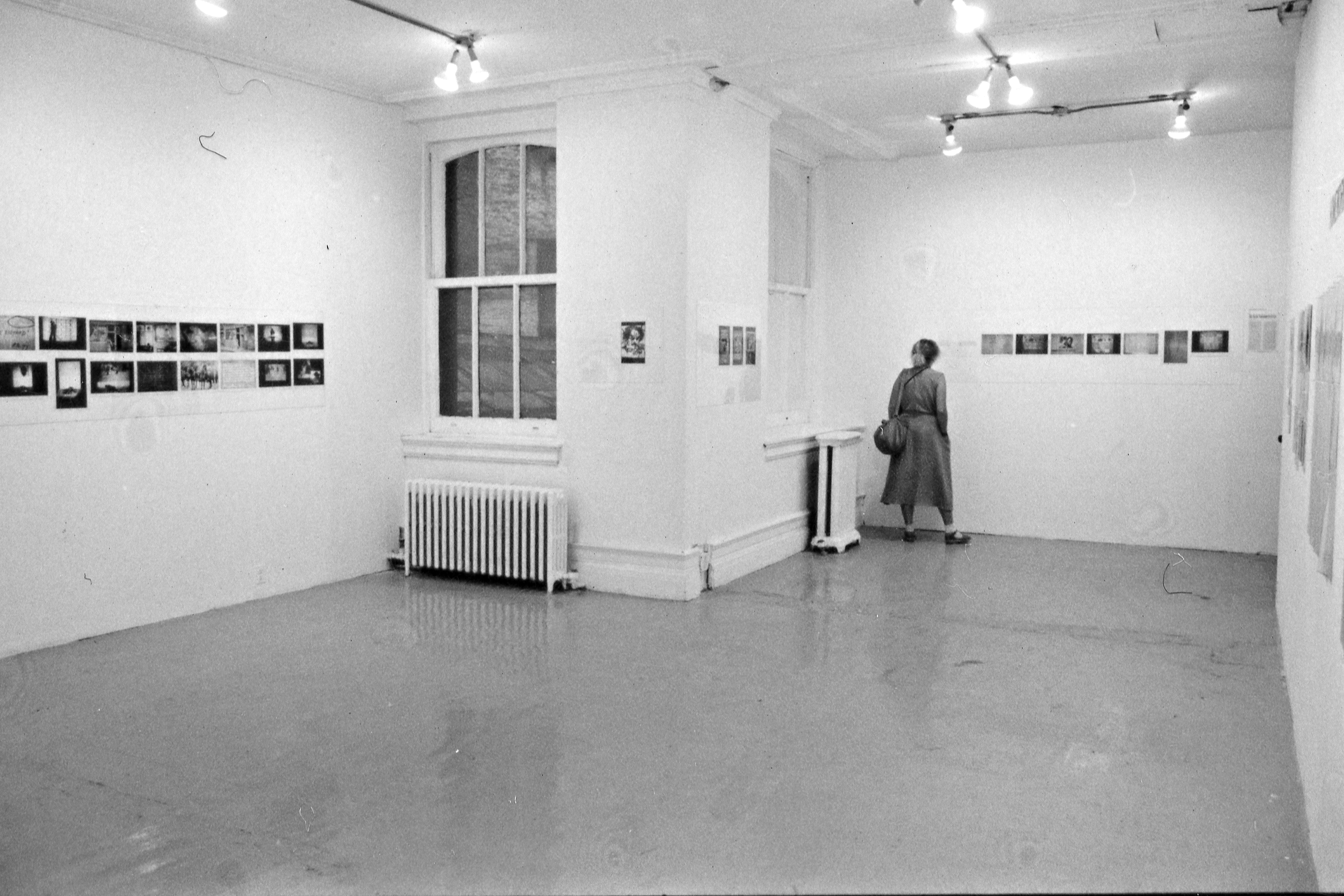 A photograph of a room with photographs and small works on paper hung on each wall. A visitor looks at the works on the far wall.