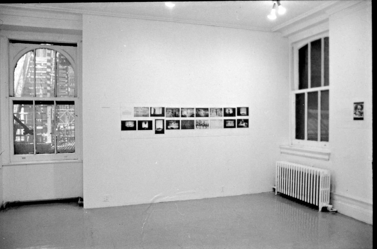 A photograph of a room with a series of photographs and works on paper hung in two rows on the far wall. A window on the left and a window on the right side are visible.