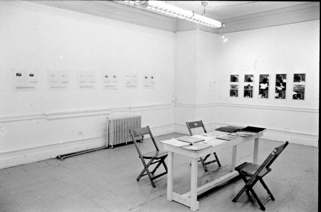A photograph of a room with works on paper hung on one wall and photographs on the other wall. In the center of the room is a table, strewn with papers, with three chairs.